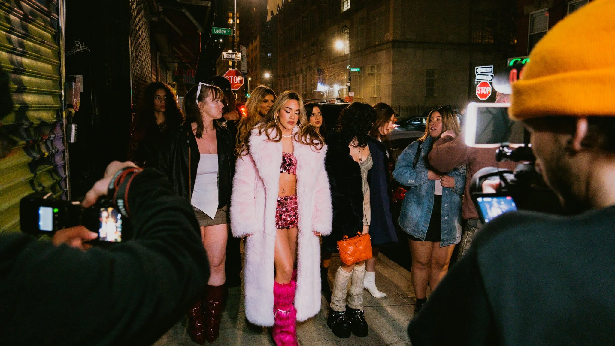 Group of young women dressed in fashionable and provocative clothing, standing on a city street at night, while being photographed and filmed by multiple cameras and phones with street signs visible in the background.