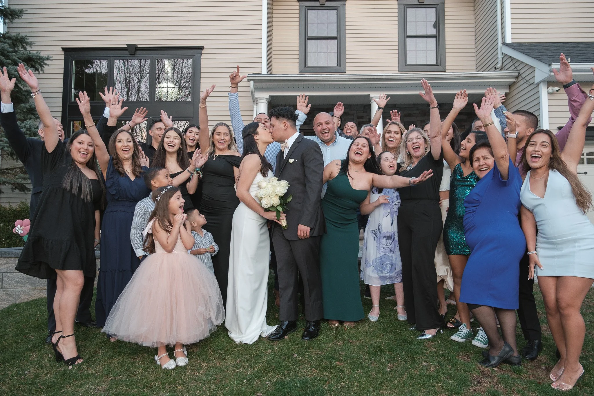A large group of people celebrating outdoors in front of a house, with a couple kissing at the center, surrounded by smiling guests.