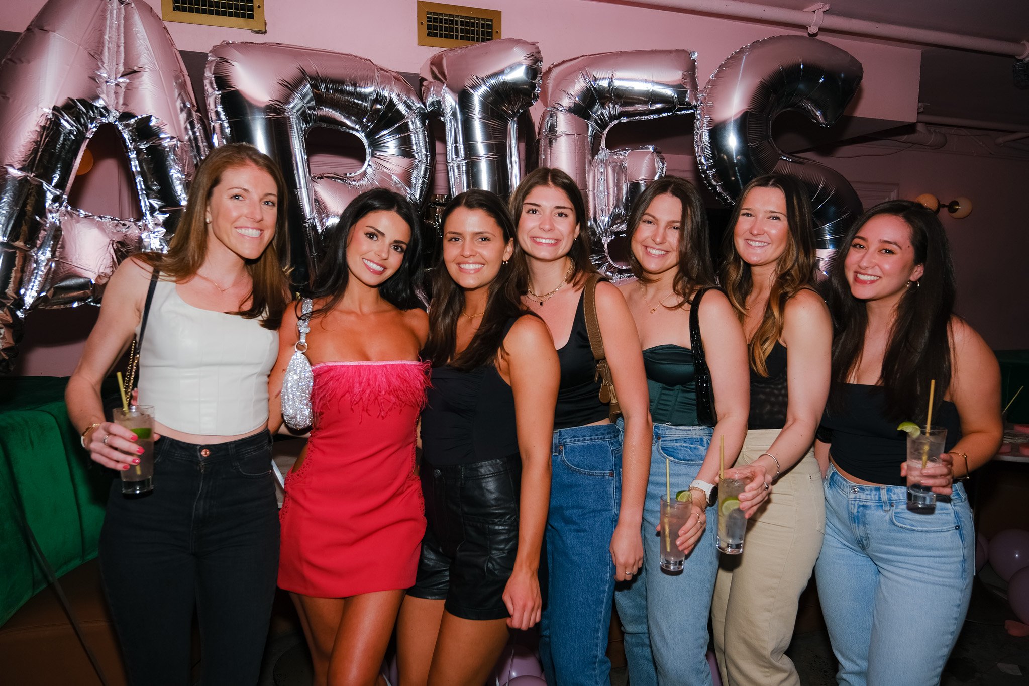 A group of seven women smiling and posing together at a party or celebration. They are standing in front of metallic balloons spelling out a word and holding drinks.