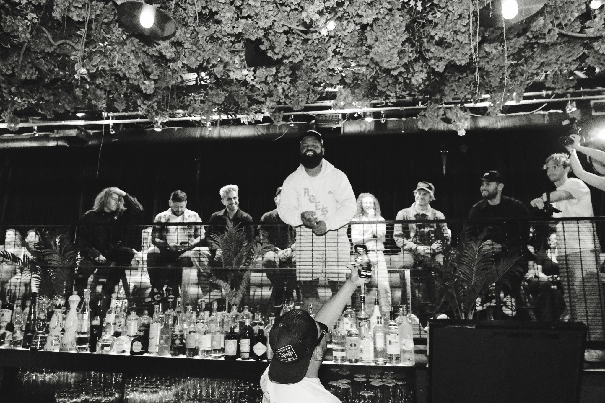 People sitting at a bar with various bottles and drinks, some standing and socializing, with a person in the foreground holding a phone, a group on an upper level enjoying the setting, decorated with hanging lights and foliage overhead, in a dimly li