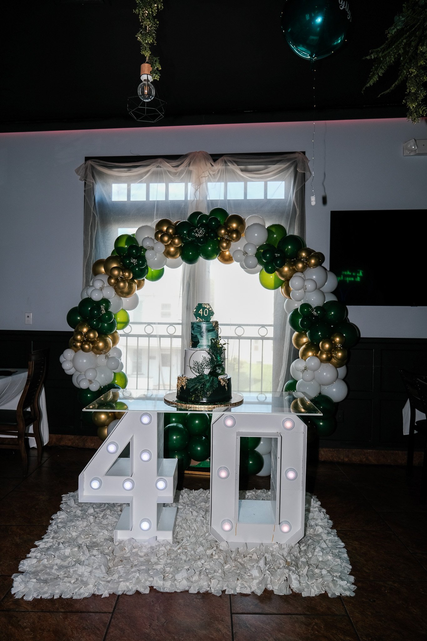 A festive celebration setup featuring a large illuminated number 40 and a balloon arch with green, white, and gold balloons. A decorated cake with the number 40 on top is placed on a table in front of a window with sheer curtains. The floor is decora