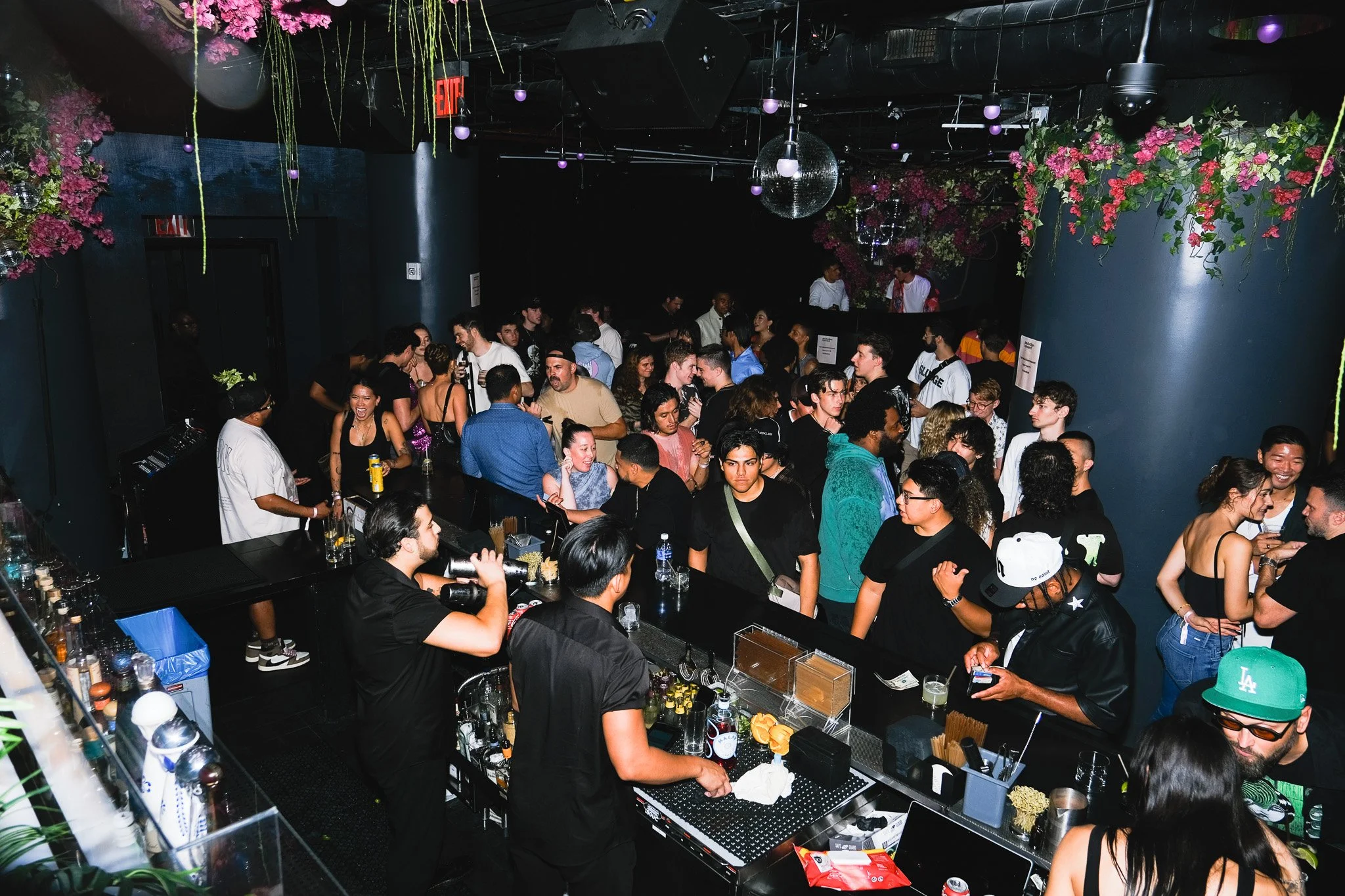 A crowded bar or club with people socializing, ordering drinks, and enjoying the nightlife. Decor includes hanging pink flowers and purple globe lights, with a disco ball overhead. There is a bartender preparing drinks in the foreground.