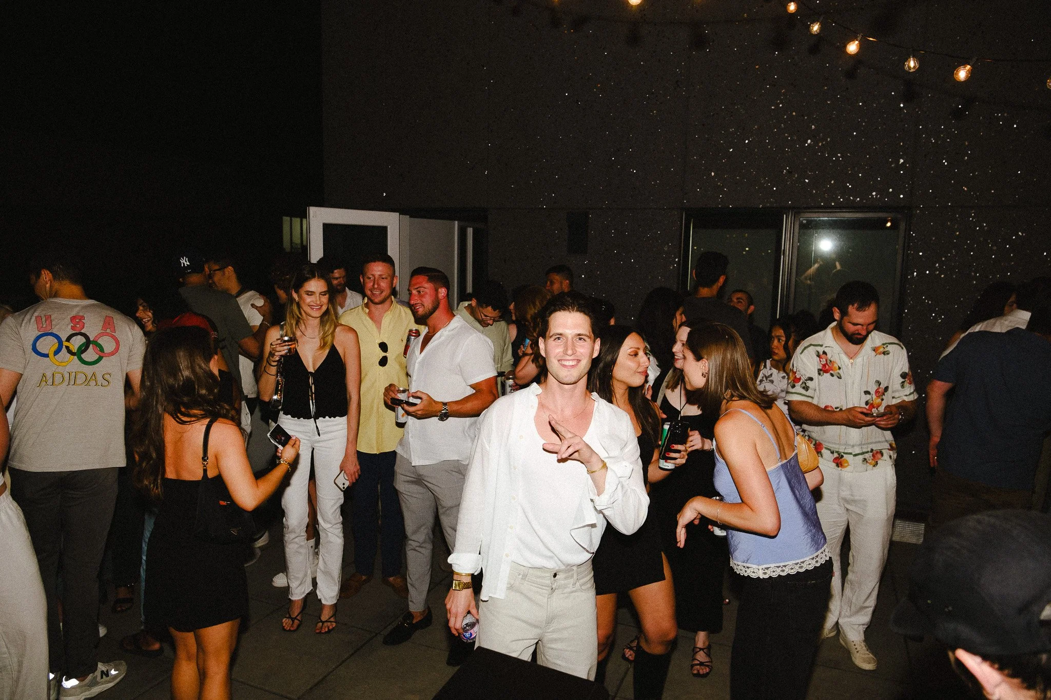 A group of people gathered at a party or social event, some holding drinks, smiling, and engaging in conversation. The setting appears to be indoors with decorative lighting on the ceiling.