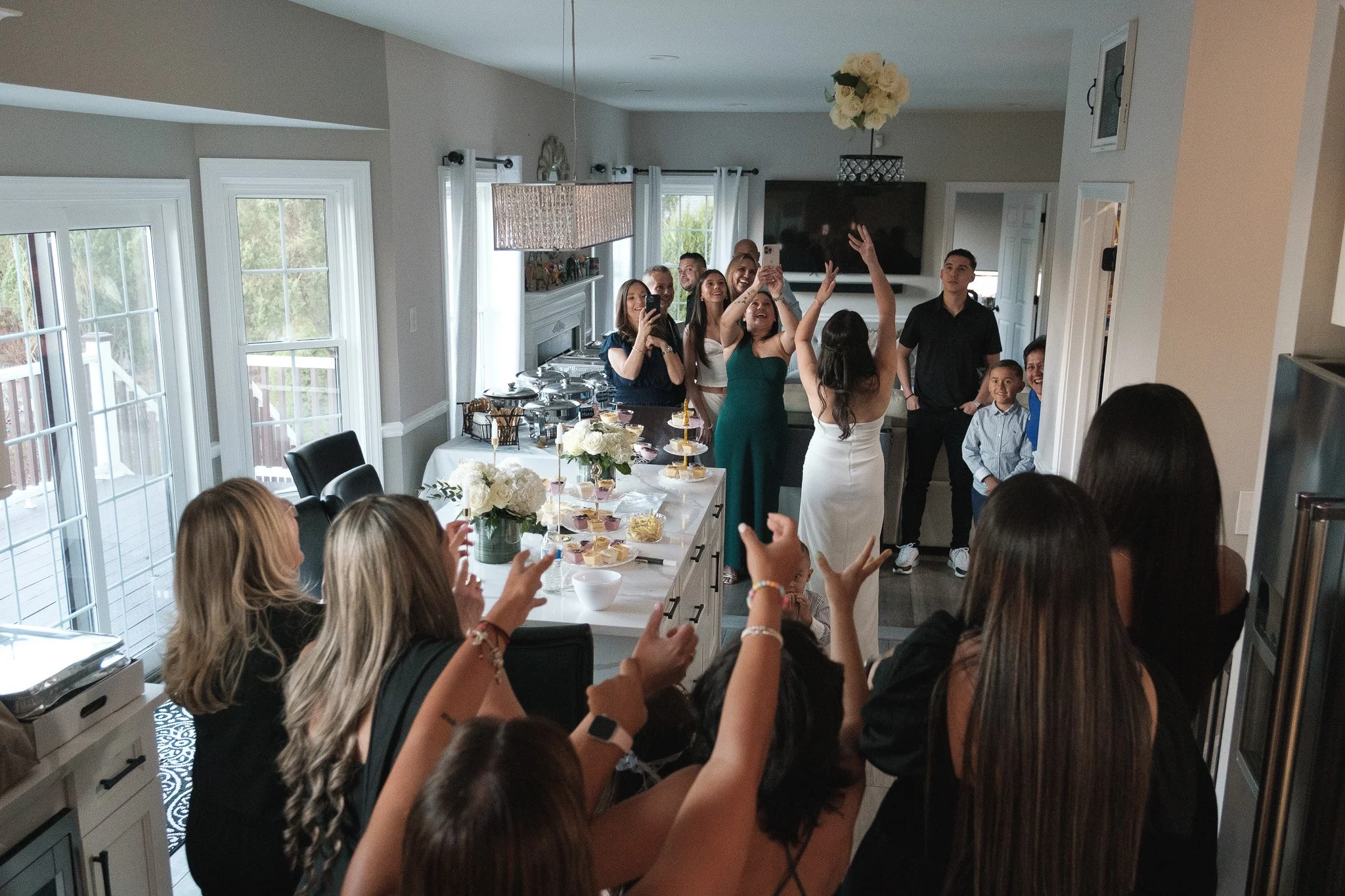 People gathered in a kitchen celebrating, with some taking photos and a table with desserts in the foreground.