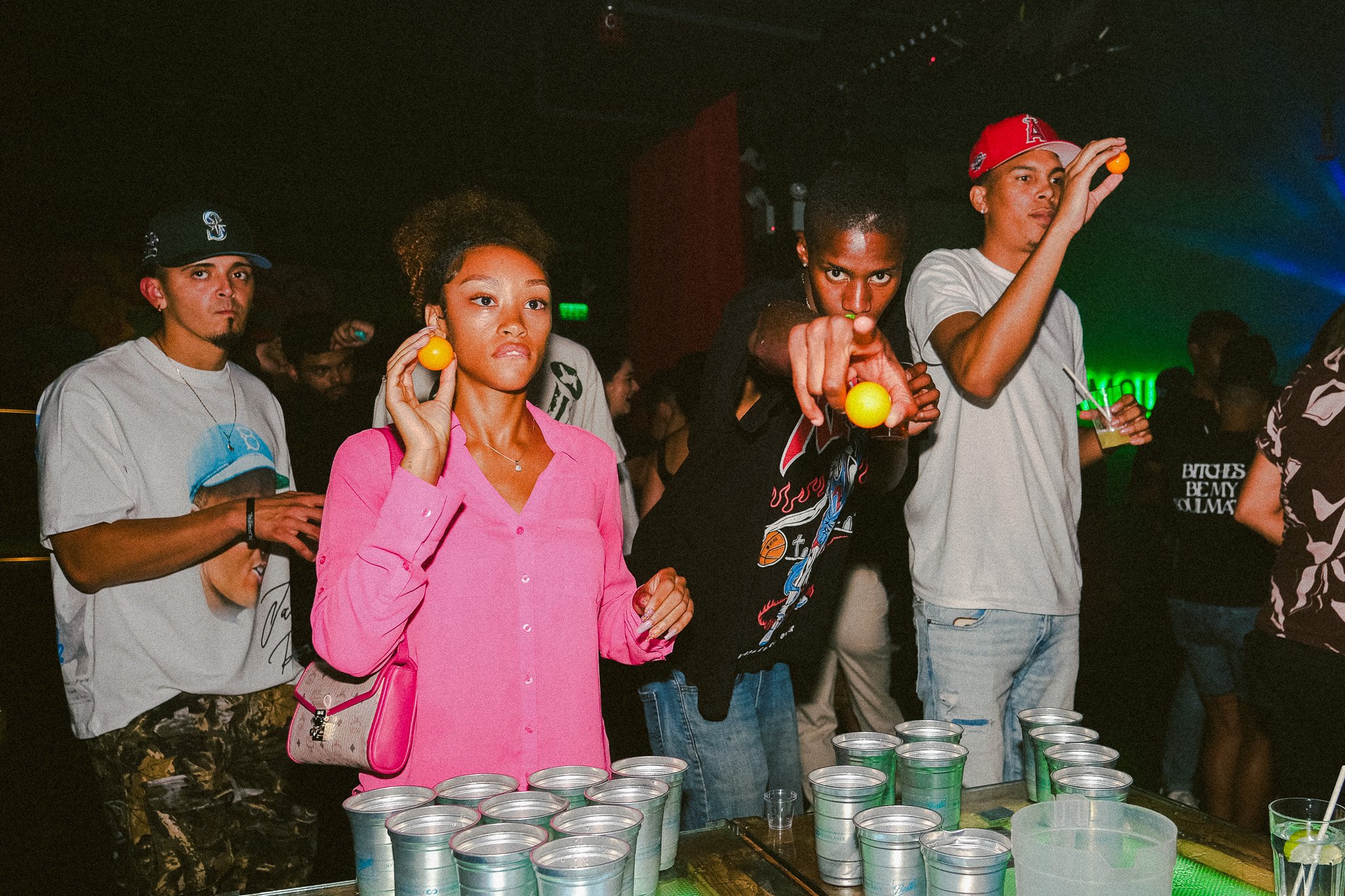 Four young adults at a party enjoying beverages. They are playing a game involving throwing small yellow balls into plastic cups on a table.