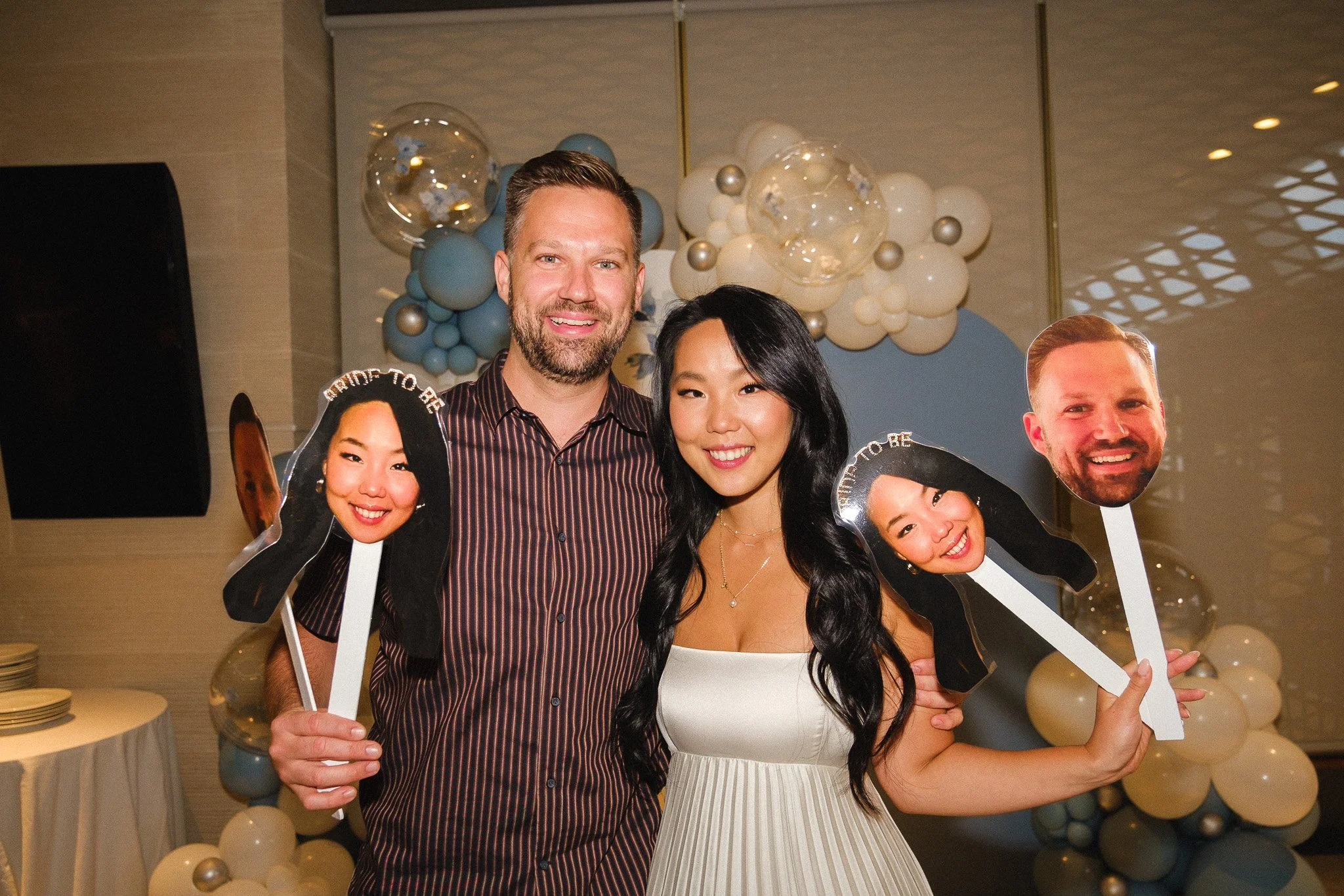 Two smiling people, a man and a woman, holding cutout photos of faces with the caption 'Proud to Be'. Background decorated with white, blue, and gold balloons.