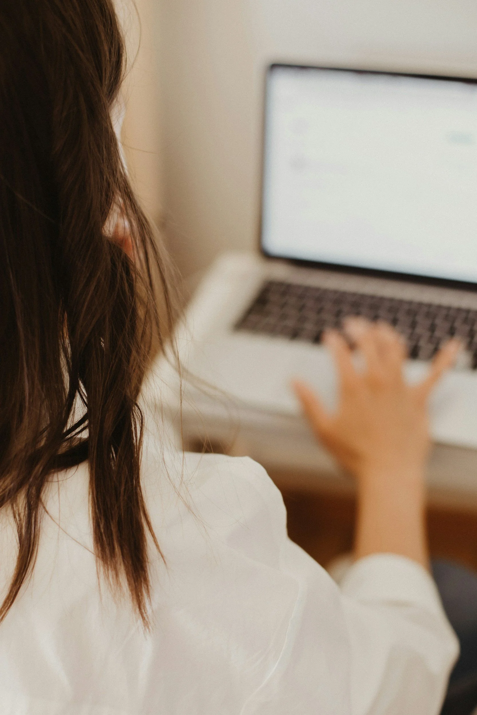 A person with long brown hair wearing a white shirt is using a laptop, with their hand on the keyboard, in an indoor setting.
