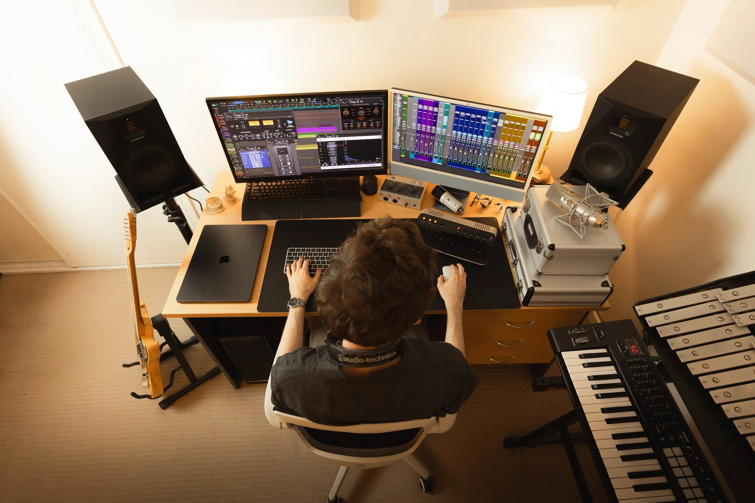 A person working at a music production studio with two large computer monitors displaying digital audio workstations, surrounded by speakers, musical instruments, and audio equipment.