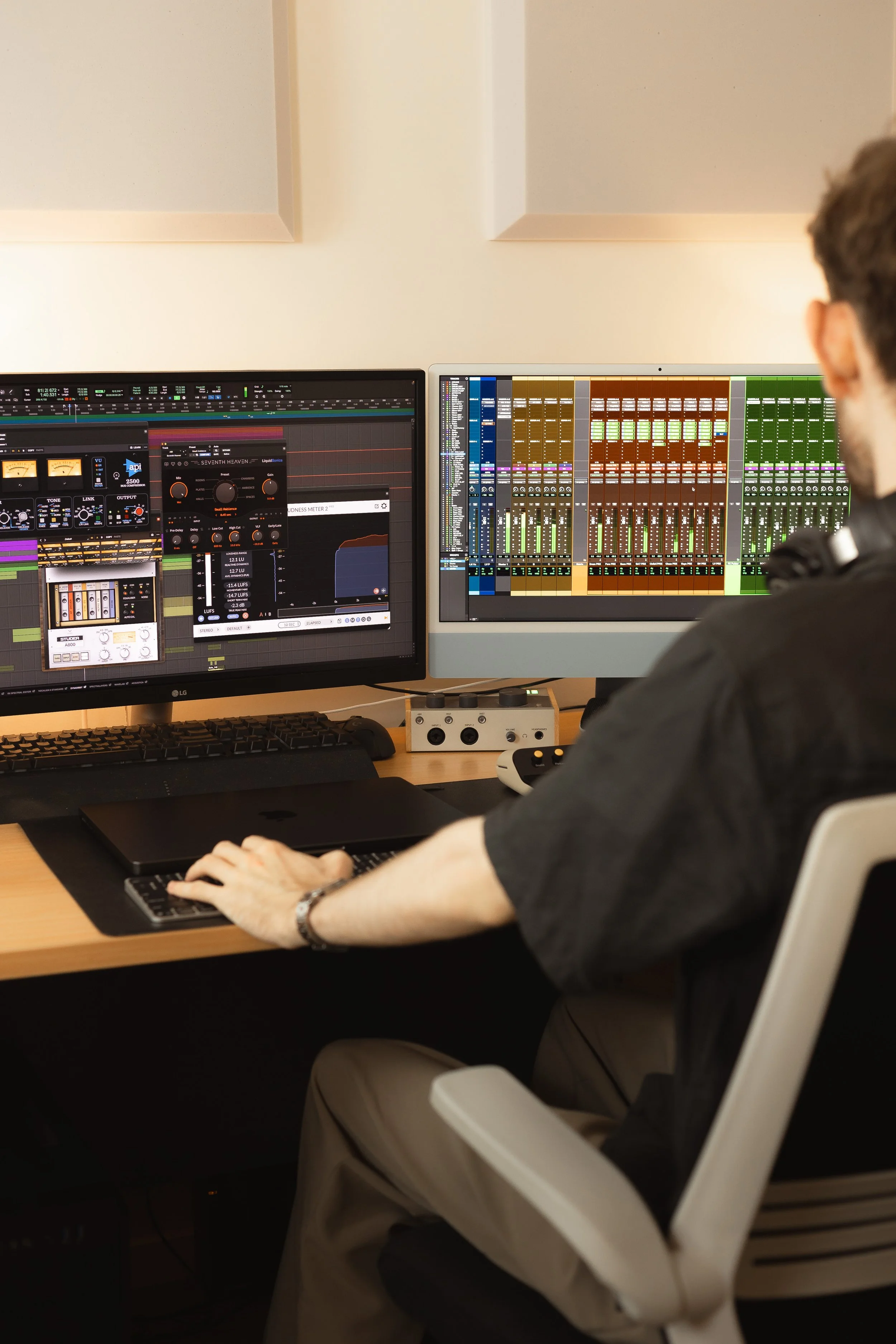 A person working on music production in a home studio, using multiple computer screens with digital audio workstations.