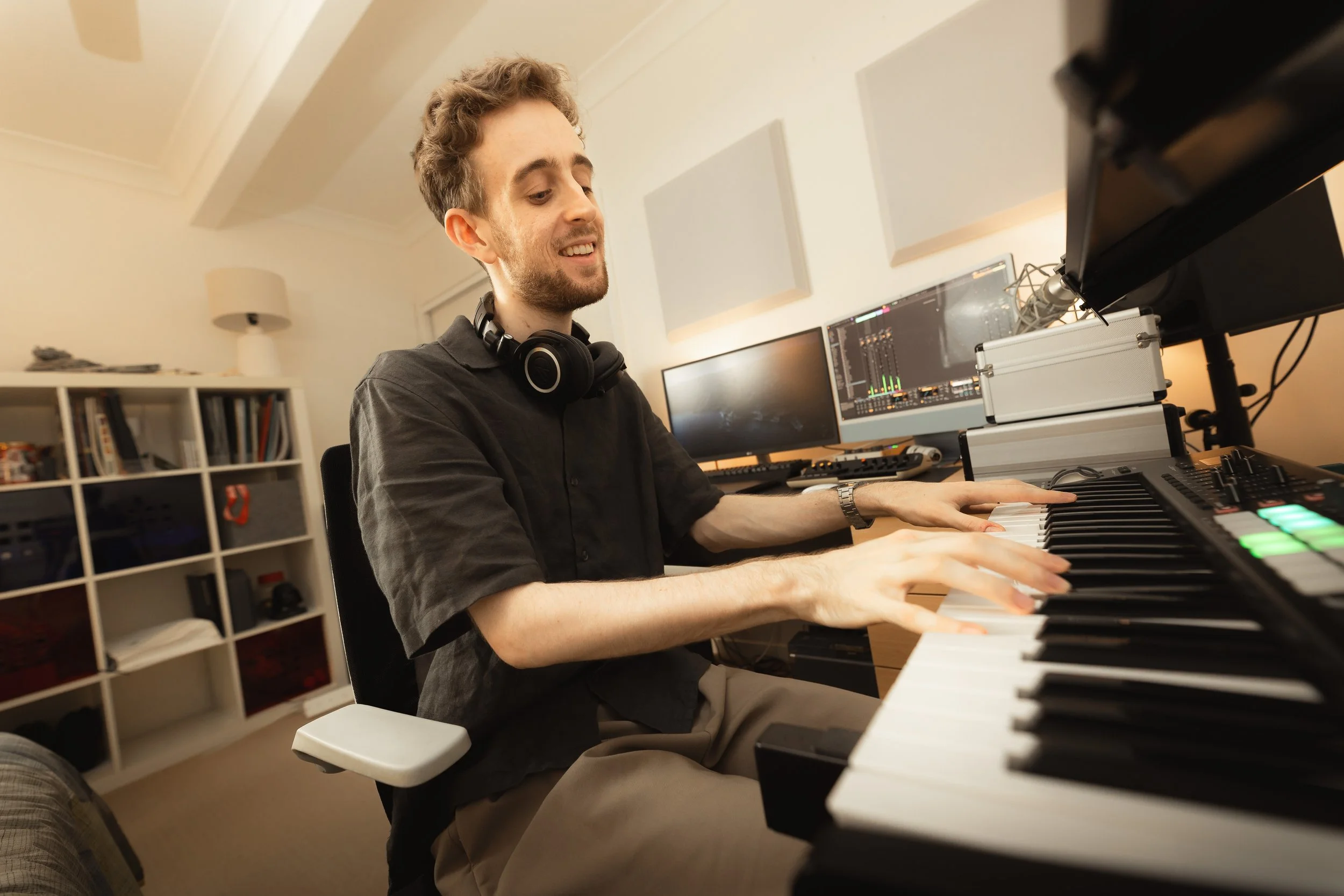 A young man with curly hair, wearing a dark shirt and headphones, playing a keyboard or digital piano in a room with musical equipment and monitors.