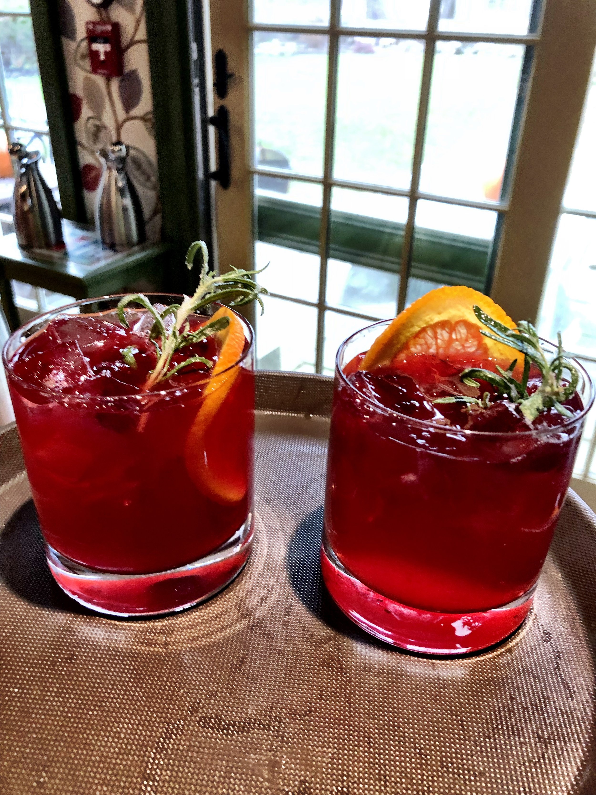 Two glasses of red cocktails with orange slices and rosemary garnishes on a tray.