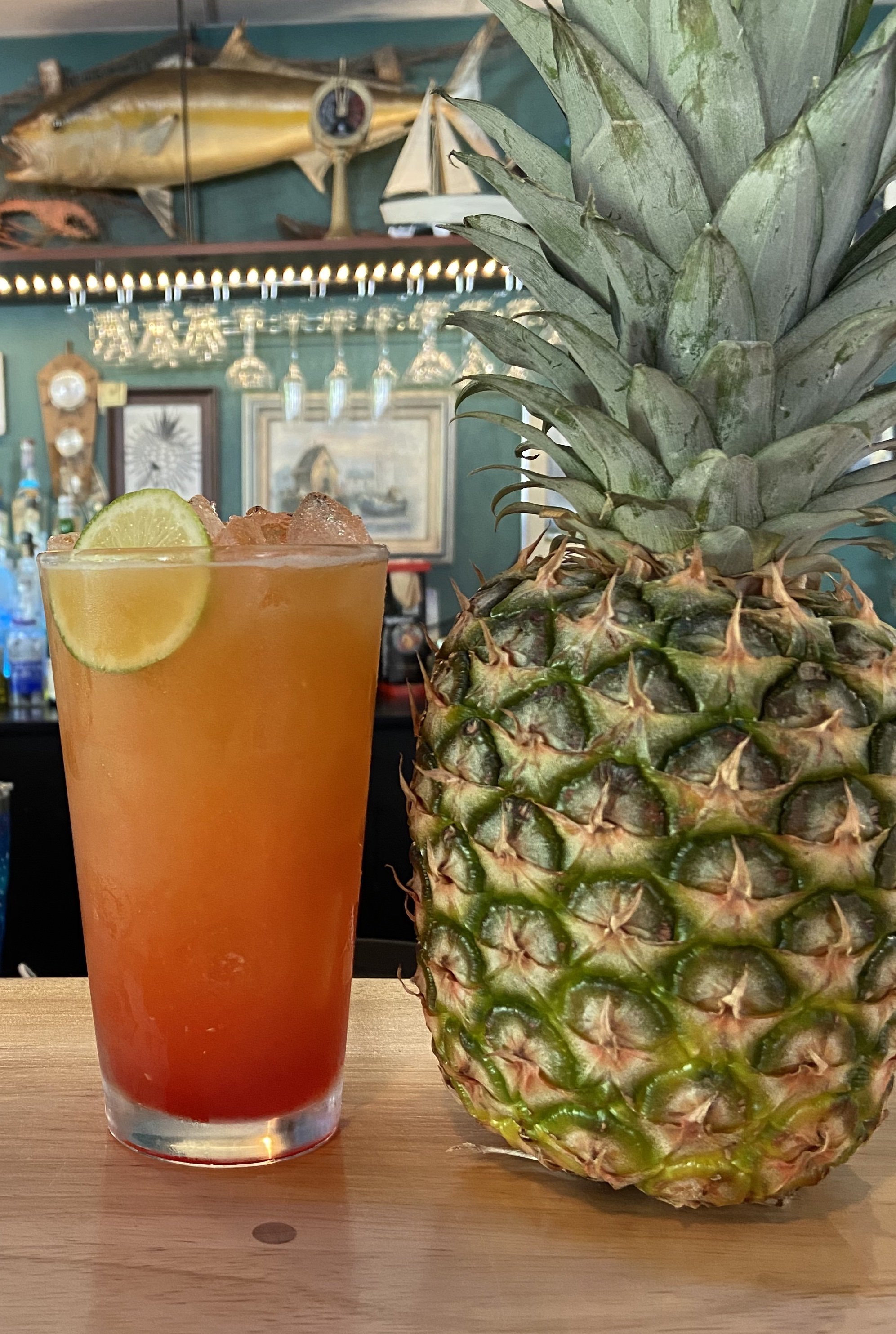 A cocktail with a lime slice next to a whole pineapple on a bar counter, with nautical decor in the background.