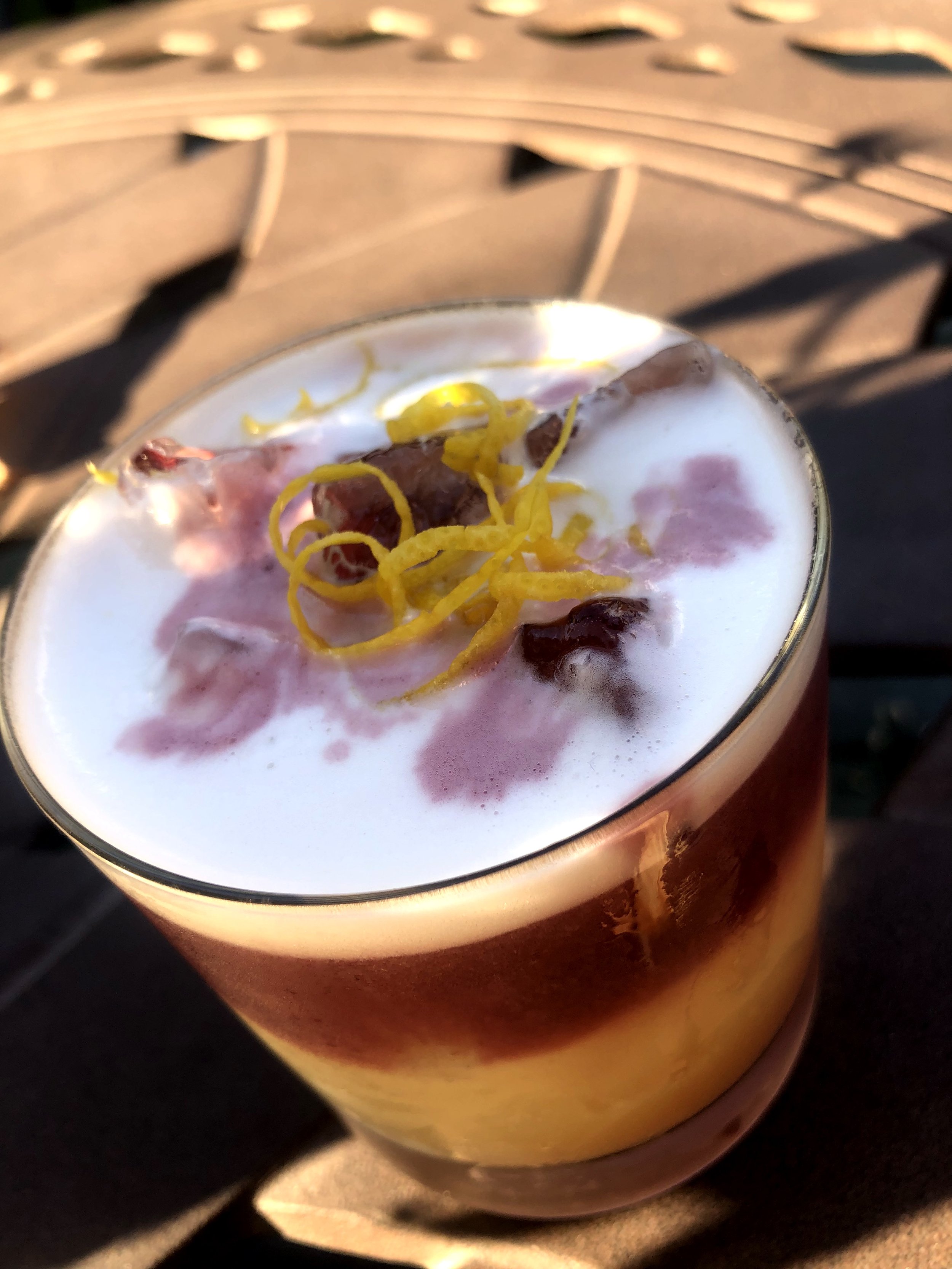 A glass of layered cocktail with foam, garnished with lemon zest and a cherry.