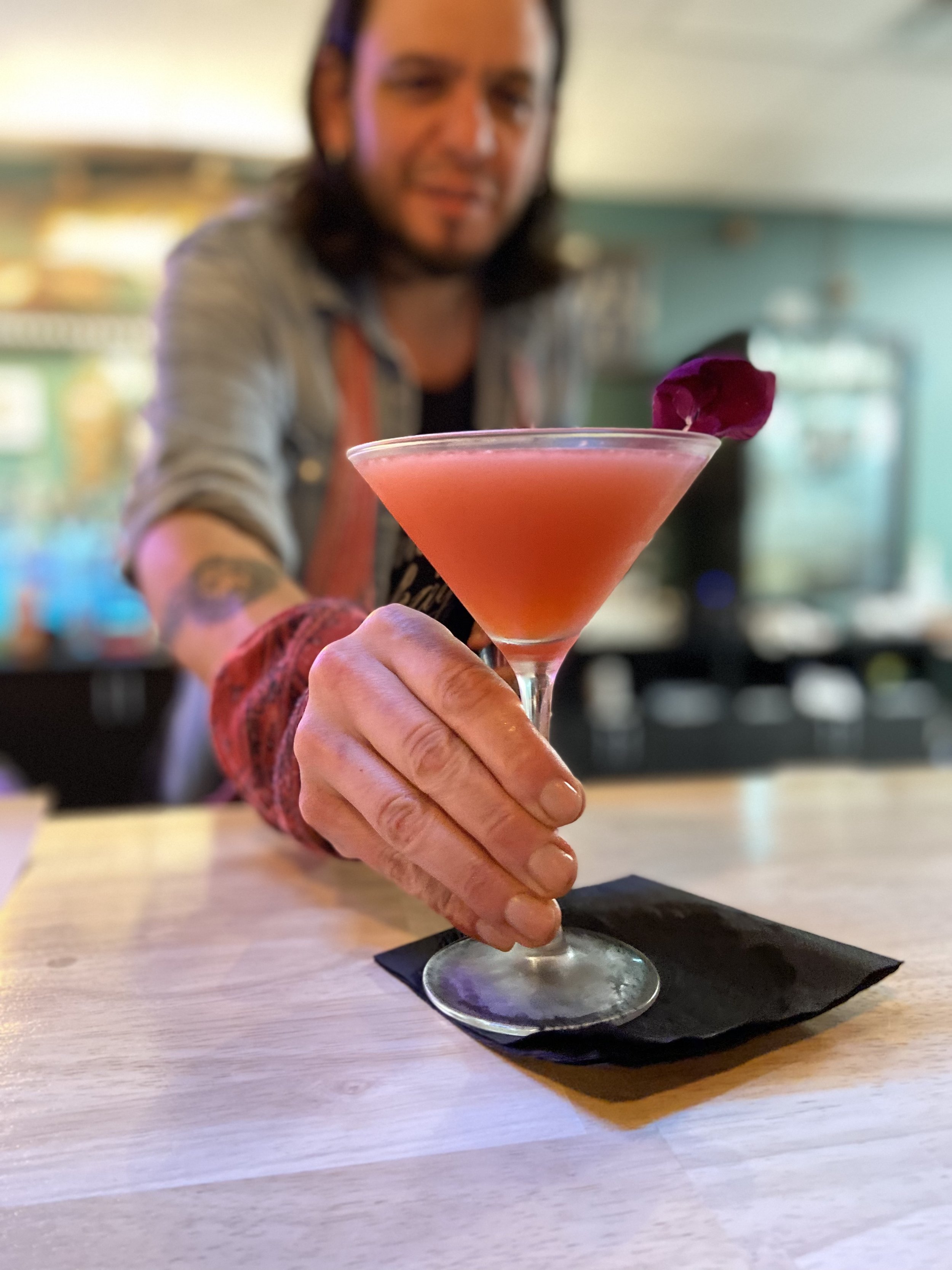 Person serving a pink cocktail in a martini glass on a black napkin, garnished with a flower.