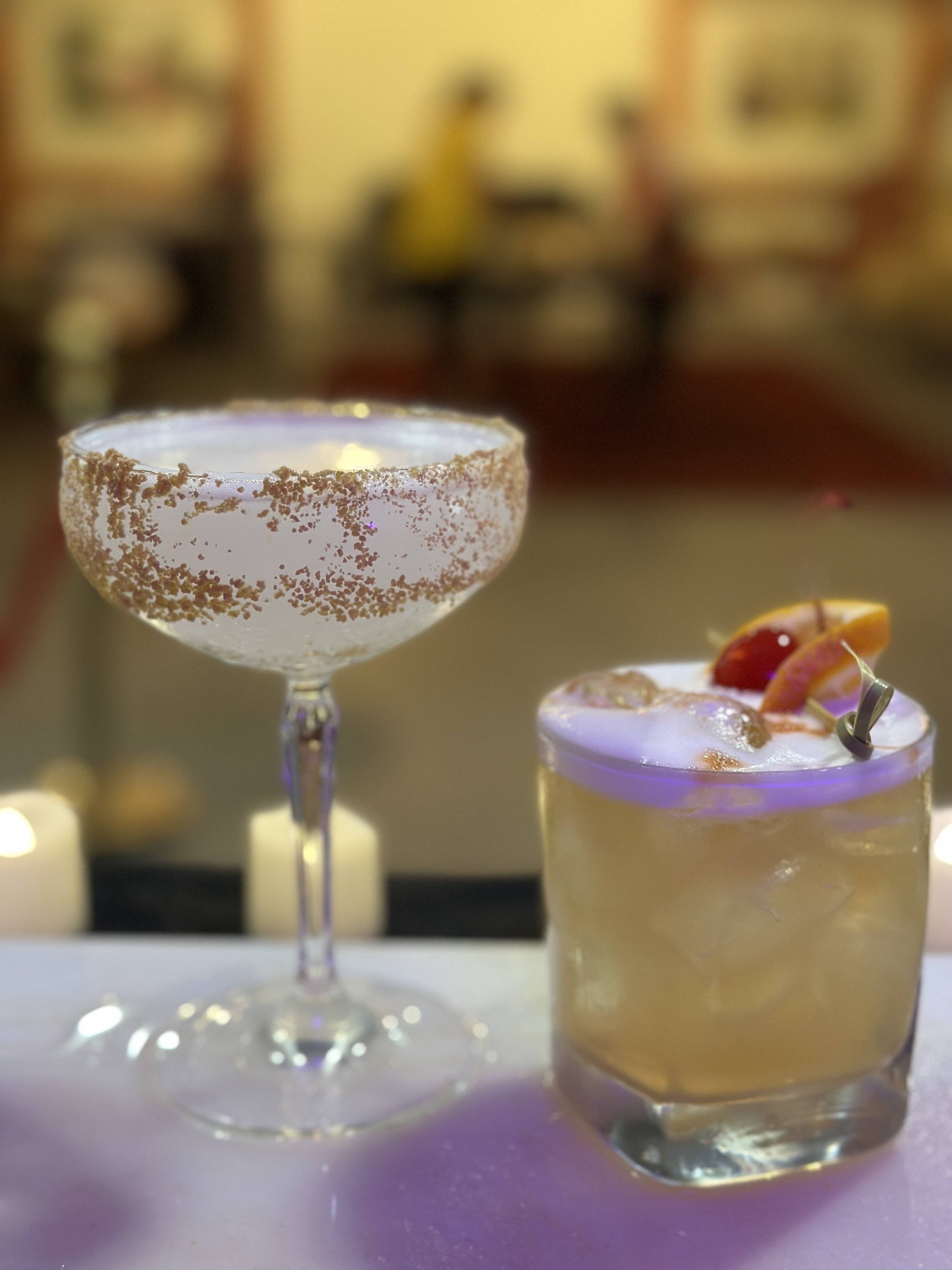 Two cocktails on a table, one in a coupe glass with a sugared rim, and the other in a rocks glass garnished with citrus slices.