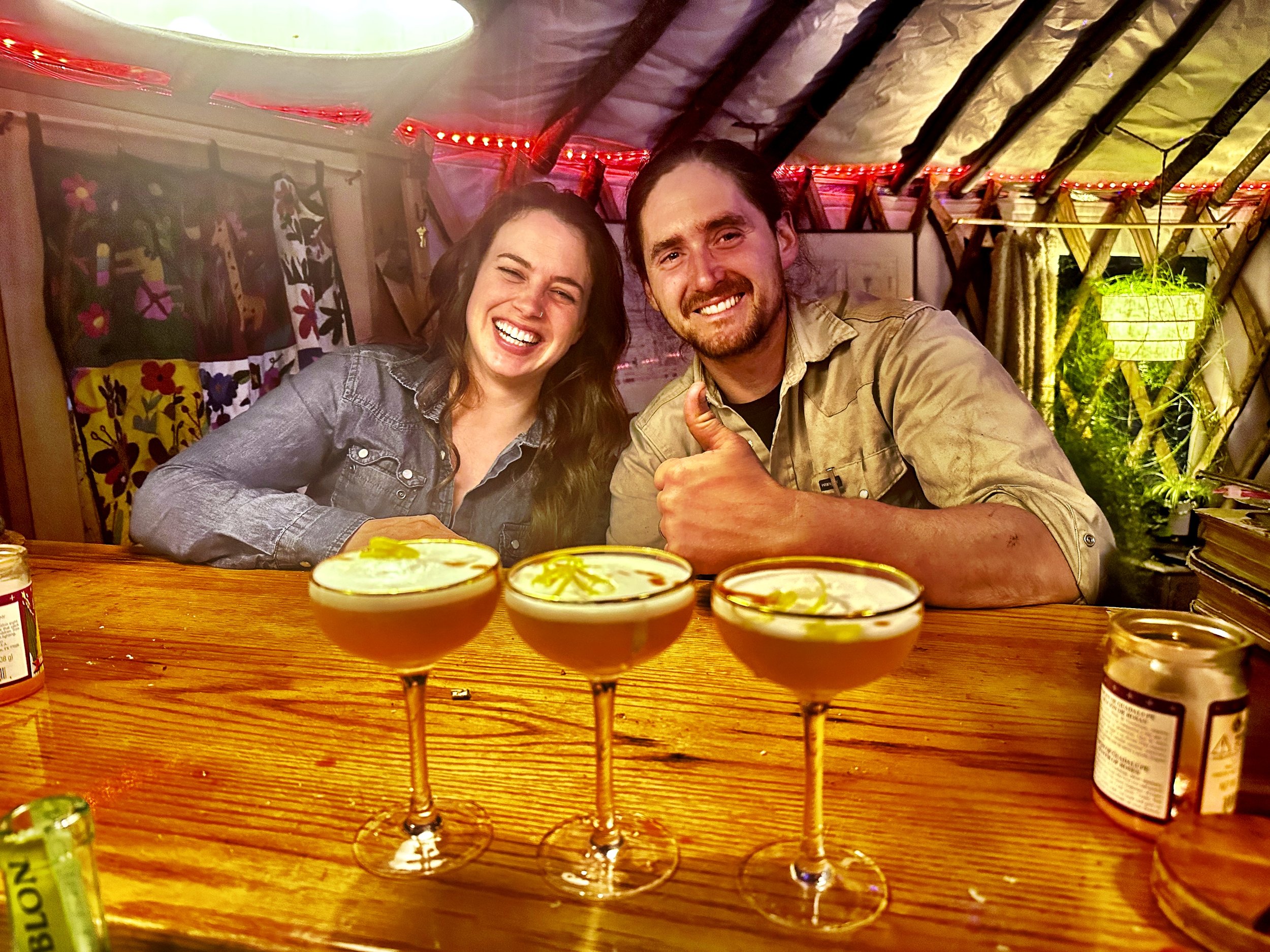 Two people smiling and sitting at a wooden bar with four cocktails in coupe glasses; the bar is inside a warmly lit, decorated space.