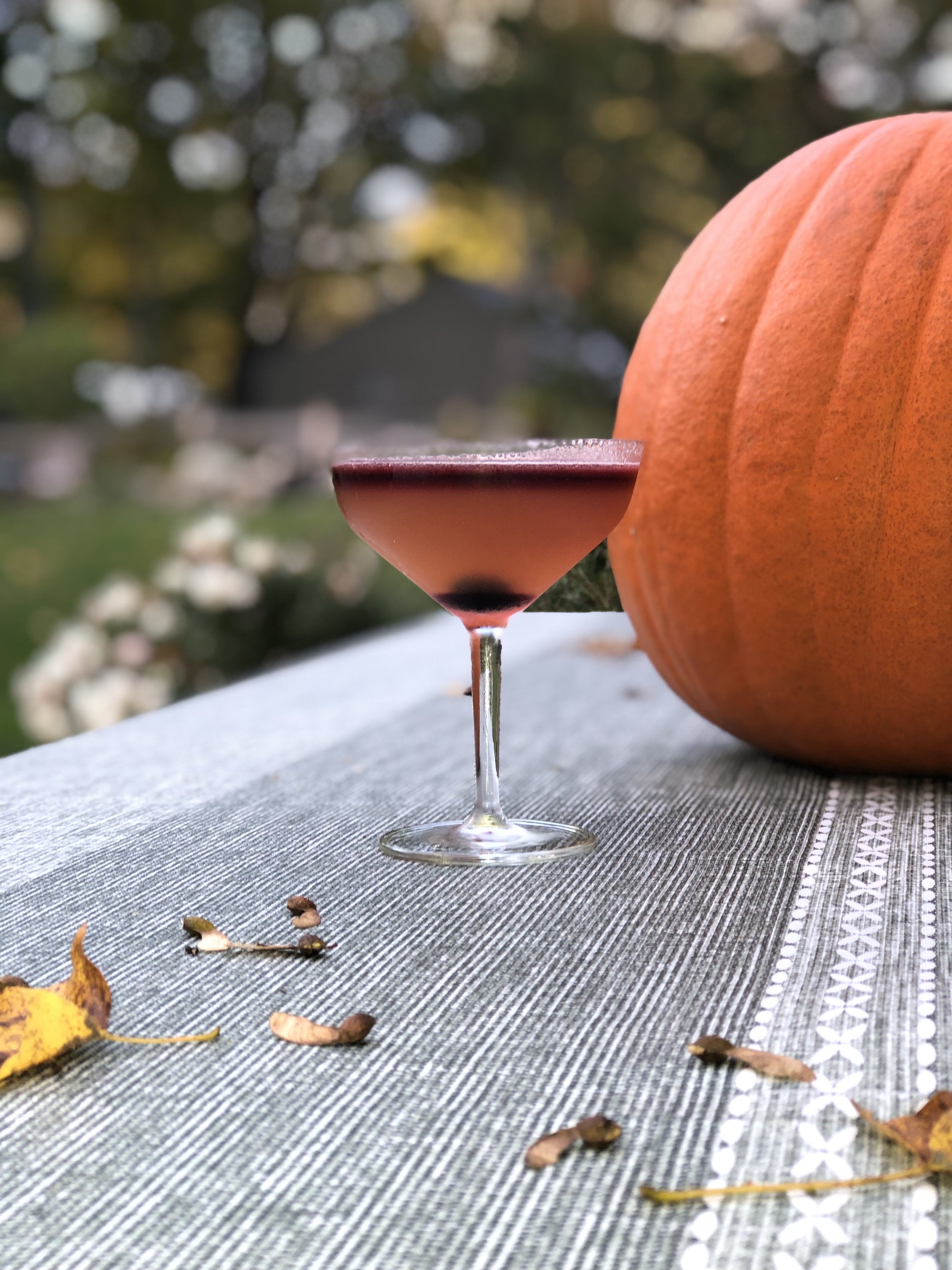 A cocktail in a glass on a table with a large pumpkin, surrounded by autumn leaves and seeds, set outdoors.