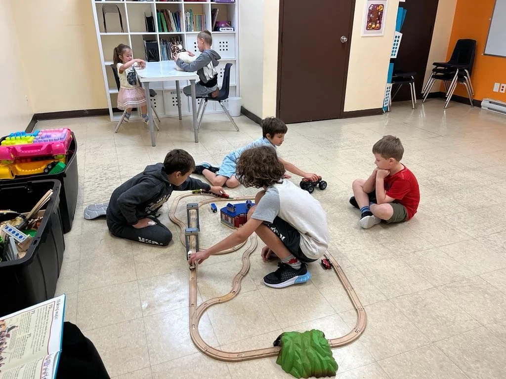 Children playing on the floor with a wooden train set, while two kids sit at a table in the background with toy dinosaurs and stuffed animals. Educational materials and toys are seen in the room.