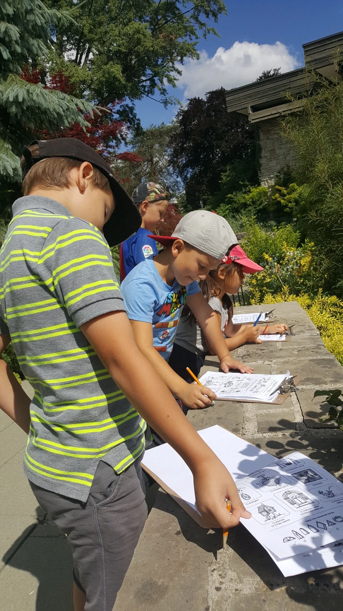 Children outdoors doing activities on clipboards, wearing casual clothes and caps, surrounded by trees and greenery.