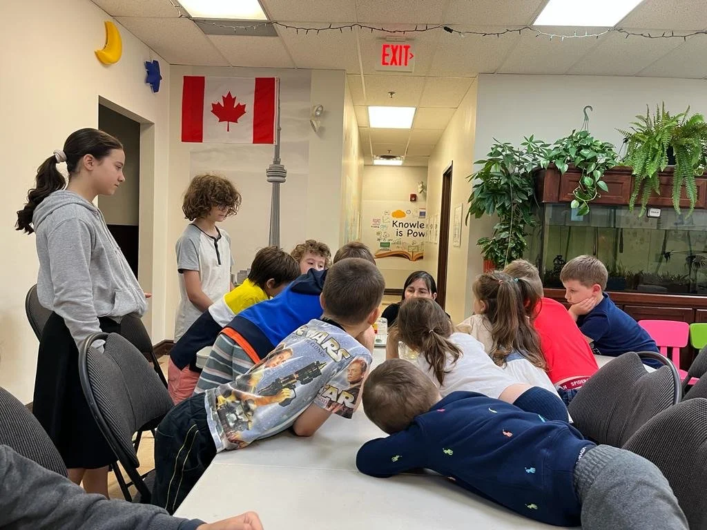 A group of children gathered around a table in a classroom setting with a Canadian flag and a CN Tower mural on the wall. The room also has potted plants and an aquarium. The children are engaged in an activity, with one standing and others sitting or leaning on the table.