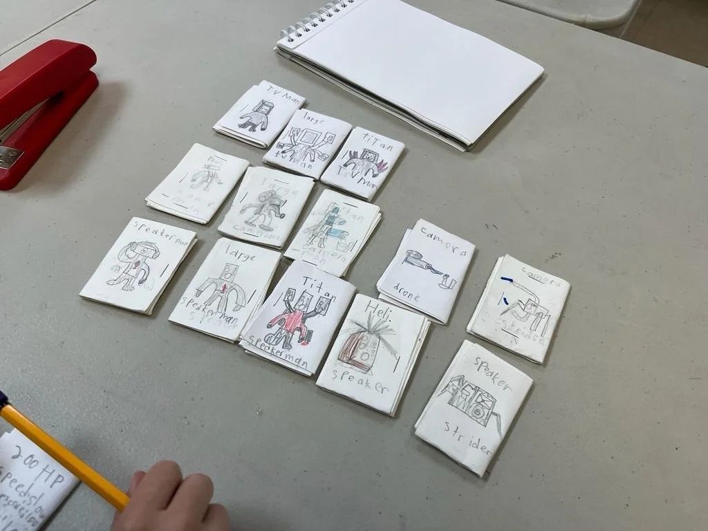 A table with handmade paper cards featuring drawings and labels of fictional characters like "Speaker Man" and "Titan." Nearby are a red stapler, a blank notebook, and a hand holding a pencil.
