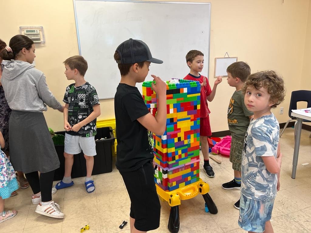 Children in a classroom building a tall stack with colorful building blocks, surrounded by various toys and supplies.