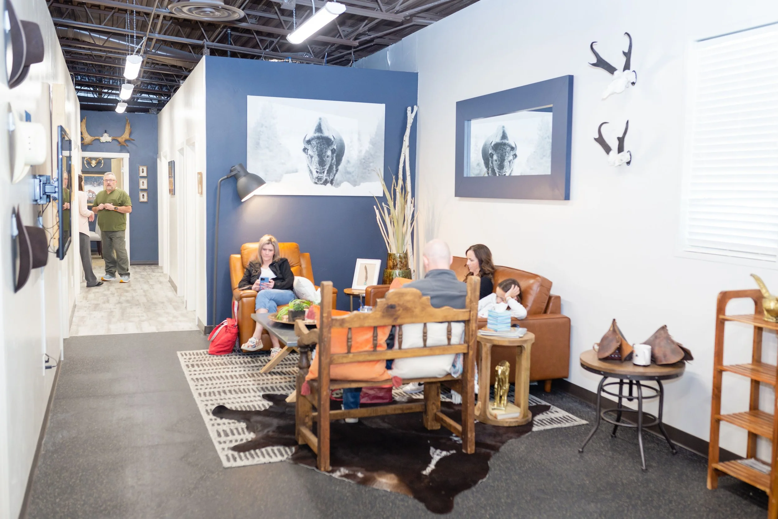 People sitting in a waiting room with leather chairs, wall art of buffalo, and antler decorations.
