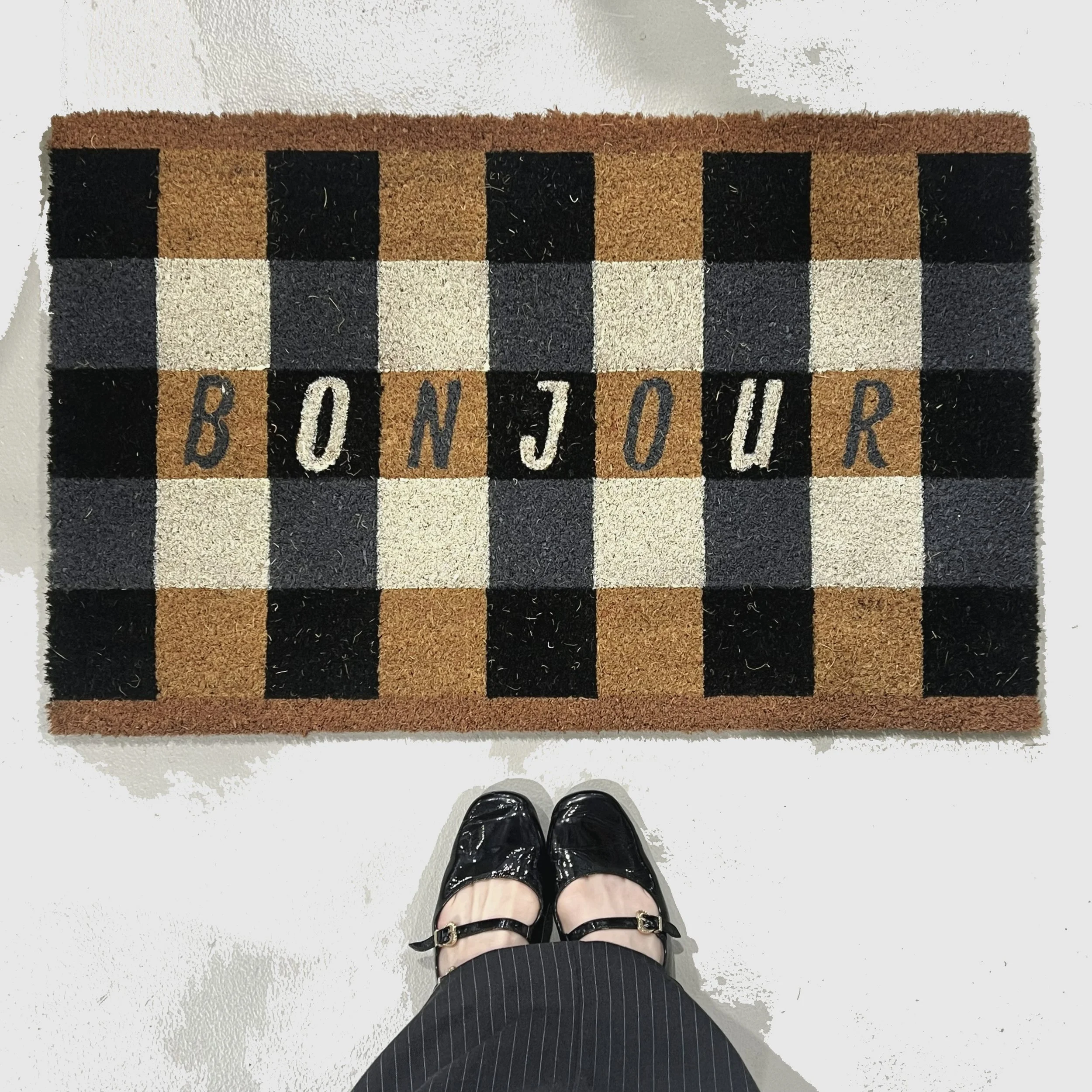 A welcome mat with a checkered pattern in black, gray, brown, and beige colors, spelling 'BONJOUR' in the center. The image is taken from above, showing a person's feet in black shiny shoes and pinstriped pants standing on a white floor.