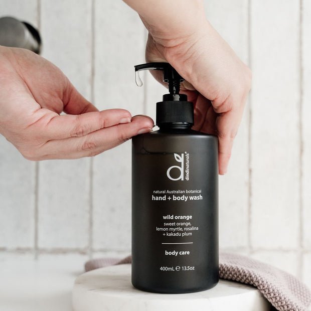 A person pressing a pump dispenser on a black bottle of Dindi Naturals hand and body wash, labeled 'wild orange' with natural ingredients like lemon myrtle and rosamarin, on a white surface with a textured cloth nearby.