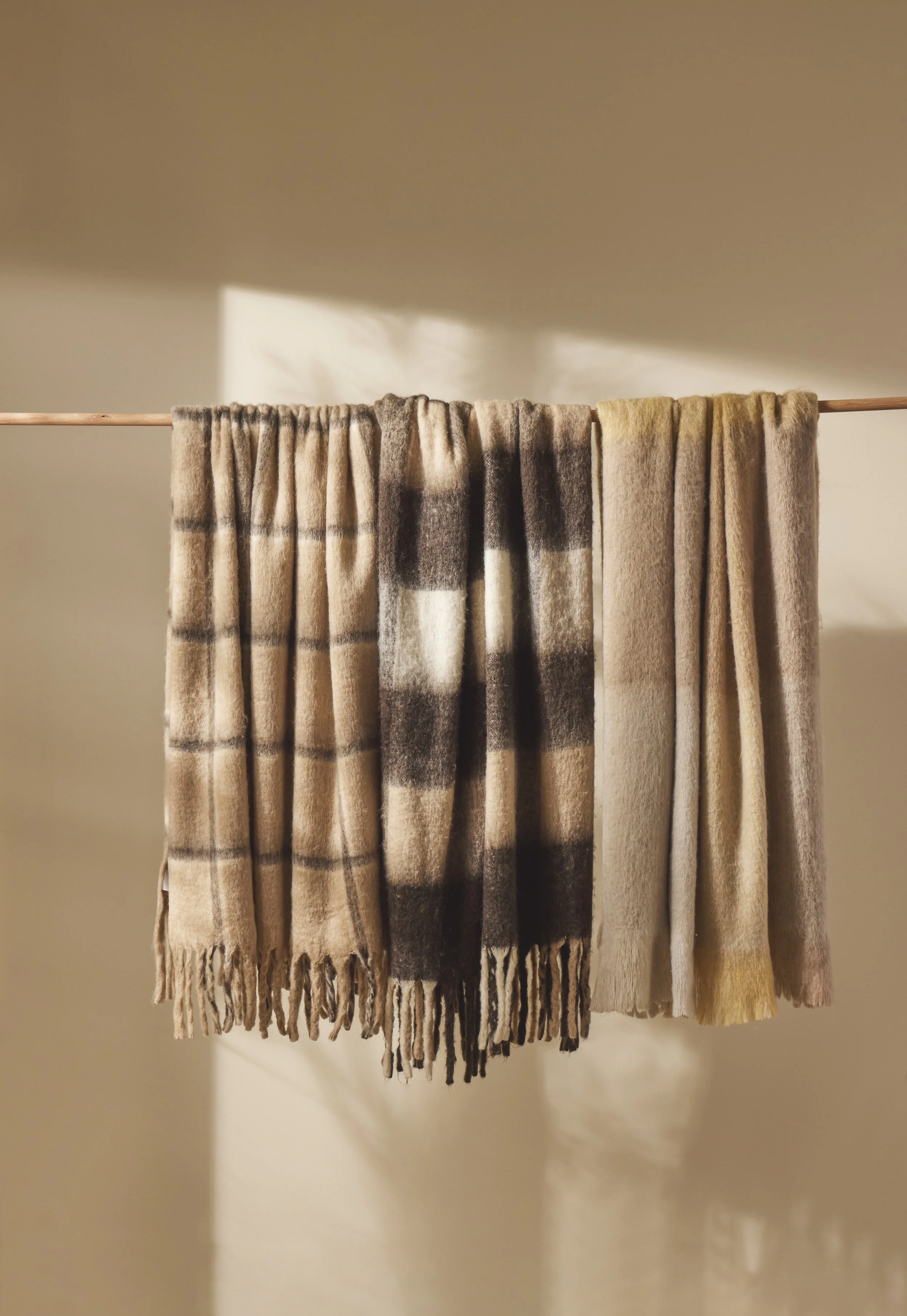 Three wool scarves hanging on a rod, with a soft beige background and shadows cast on the wall.