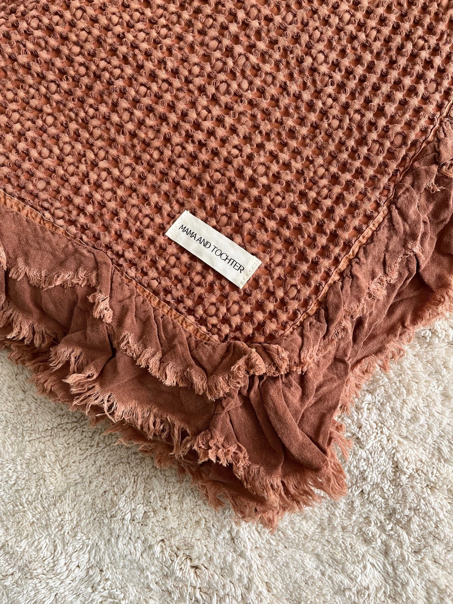 A pink textured blanket with frayed edges and a black and white "MAMA AND TOCHTER" label, placed on a white shaggy rug.