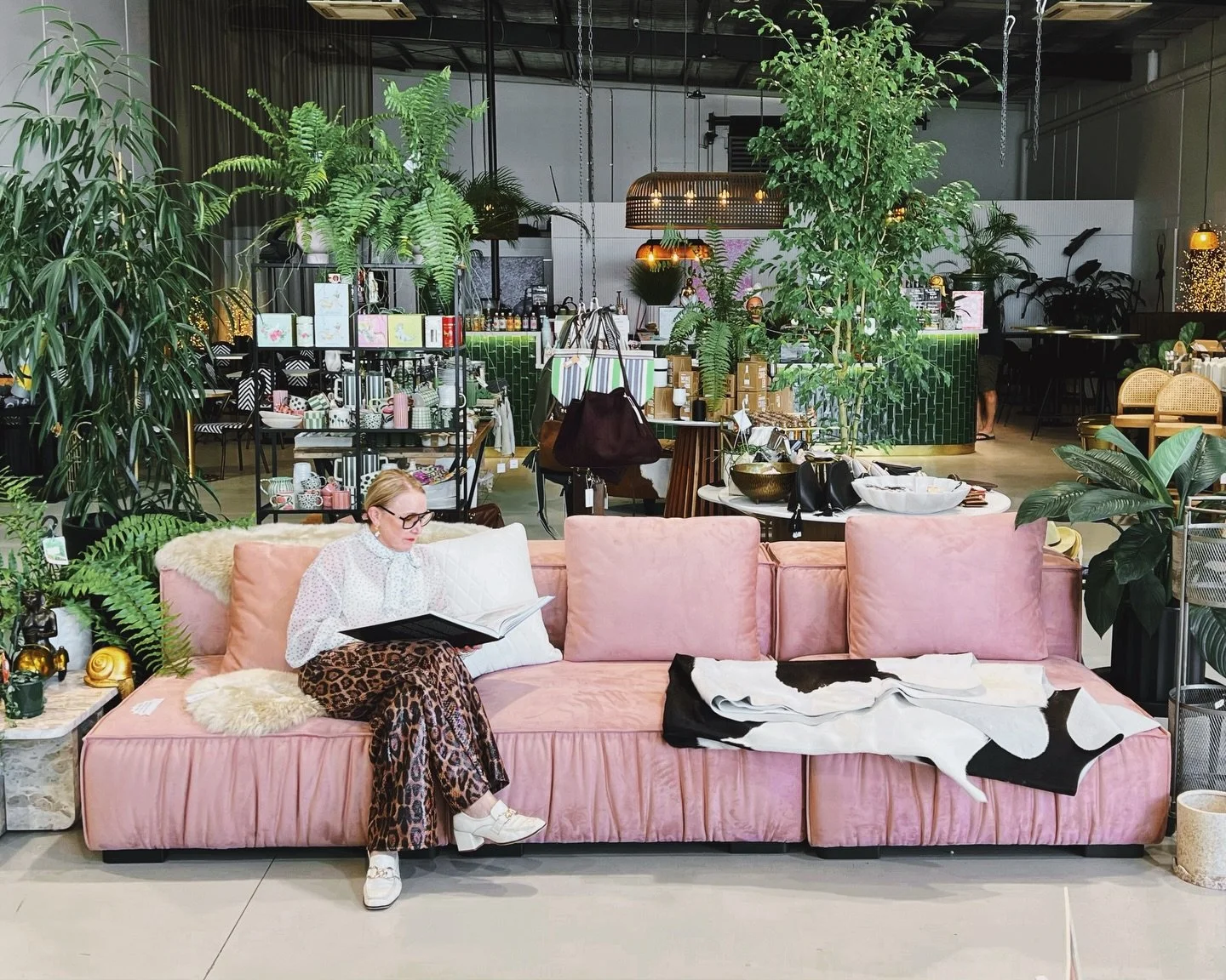 A woman with glasses and leopard print pants sitting on a pink velvet sofa in a store or cafe, reading a menu or book, with various plants and merchandise around her.
