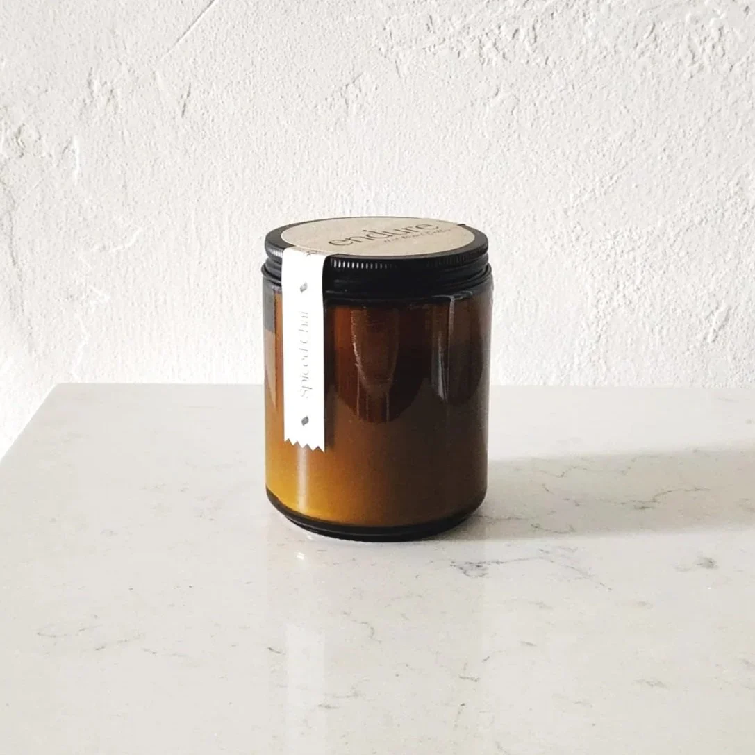 An amber glass candle in the scent spiced chair on a white surface and a white textured wall in the background.