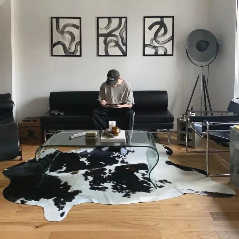 A person sitting on a black sofa in a modern living room, looking at a tablet. The room has a cowhide rug on the wooden floor, a glass coffee table in front, three framed abstract art pieces on the wall, a large floor lamp to the right, and a black armchair to the left.