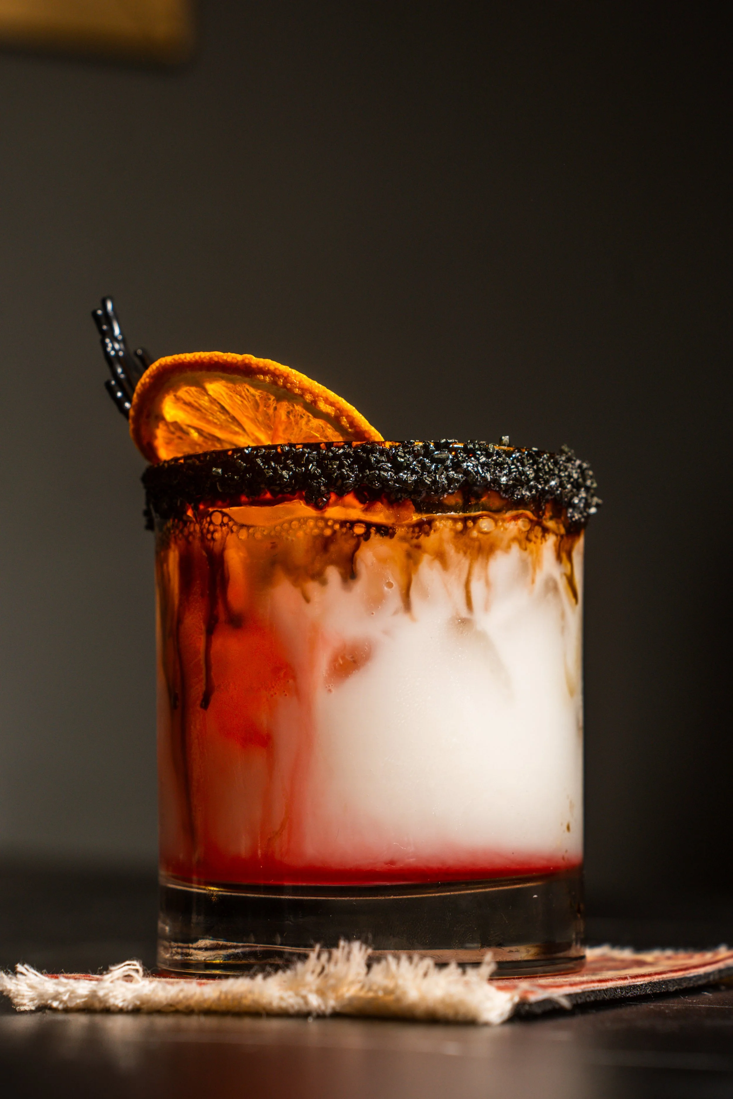 A cocktail drink in a glass with a salted rim, garnished with a dried orange slice, on a dark surface.