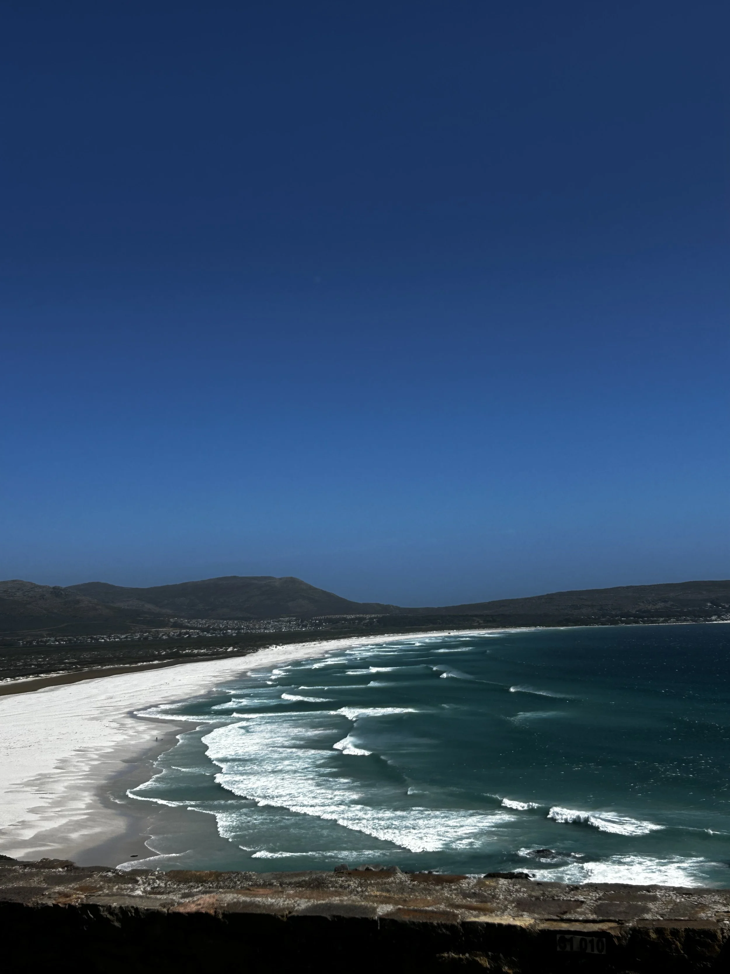 Aerial view of a beach with white sand, turquoise waves, under a clear blue sky, with mountains in the background.