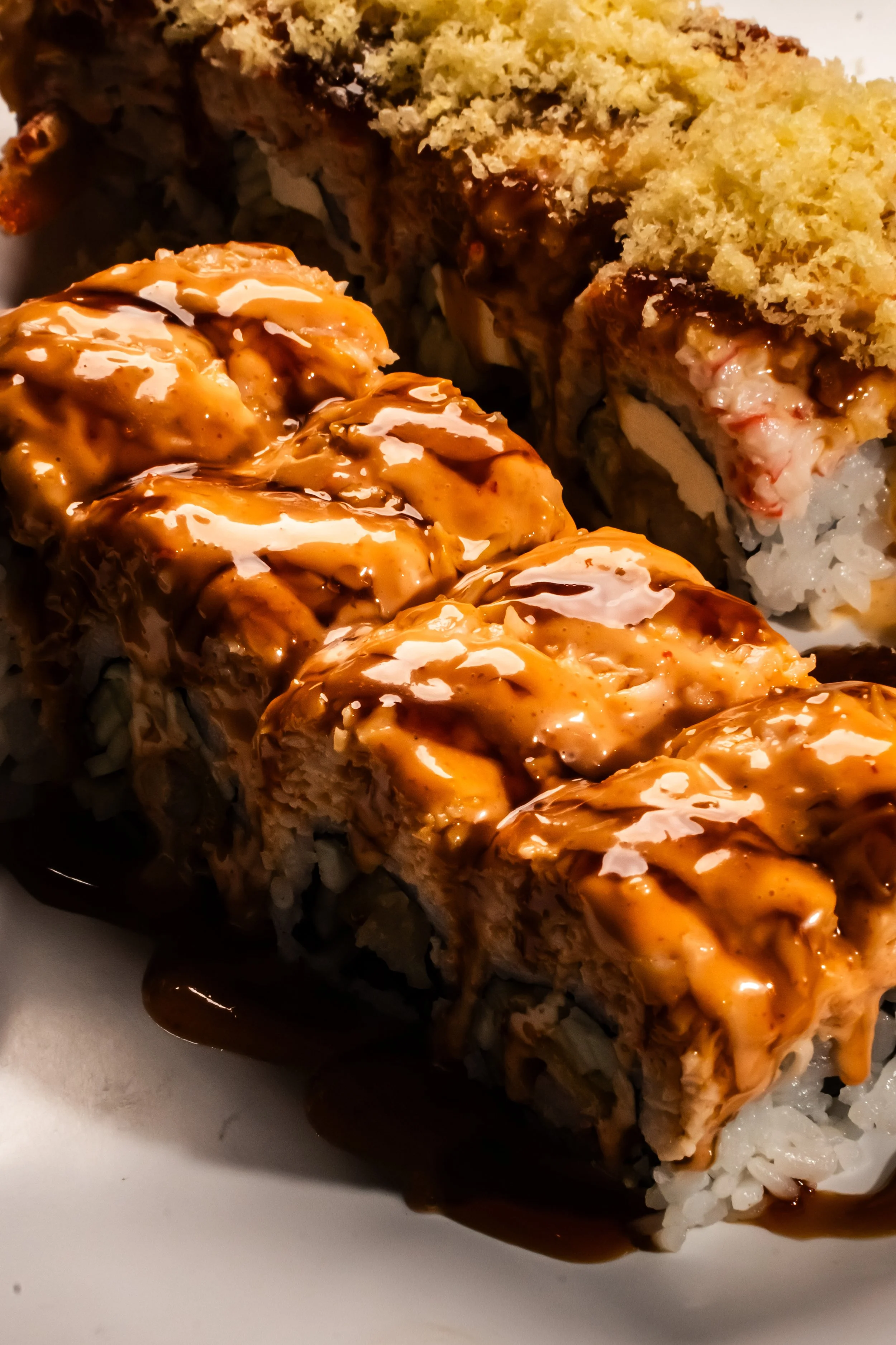 Close-up of sushi rolls with orange sauce and dark sauce drizzled on top, served on a white plate.