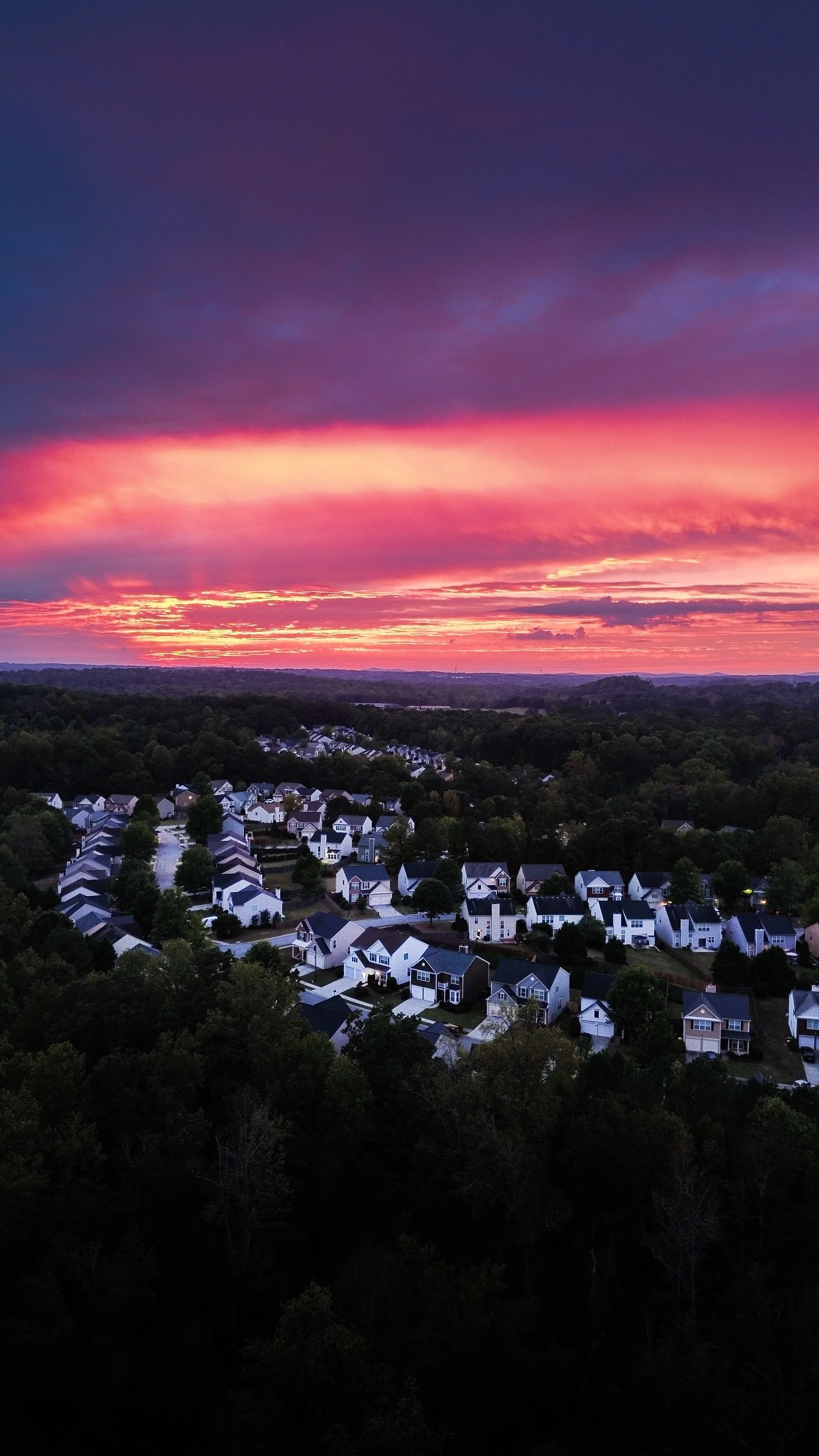 Aerial view of suburban neighborhood during sunset with houses, trees, and a colorful sky with pink and purple hues.