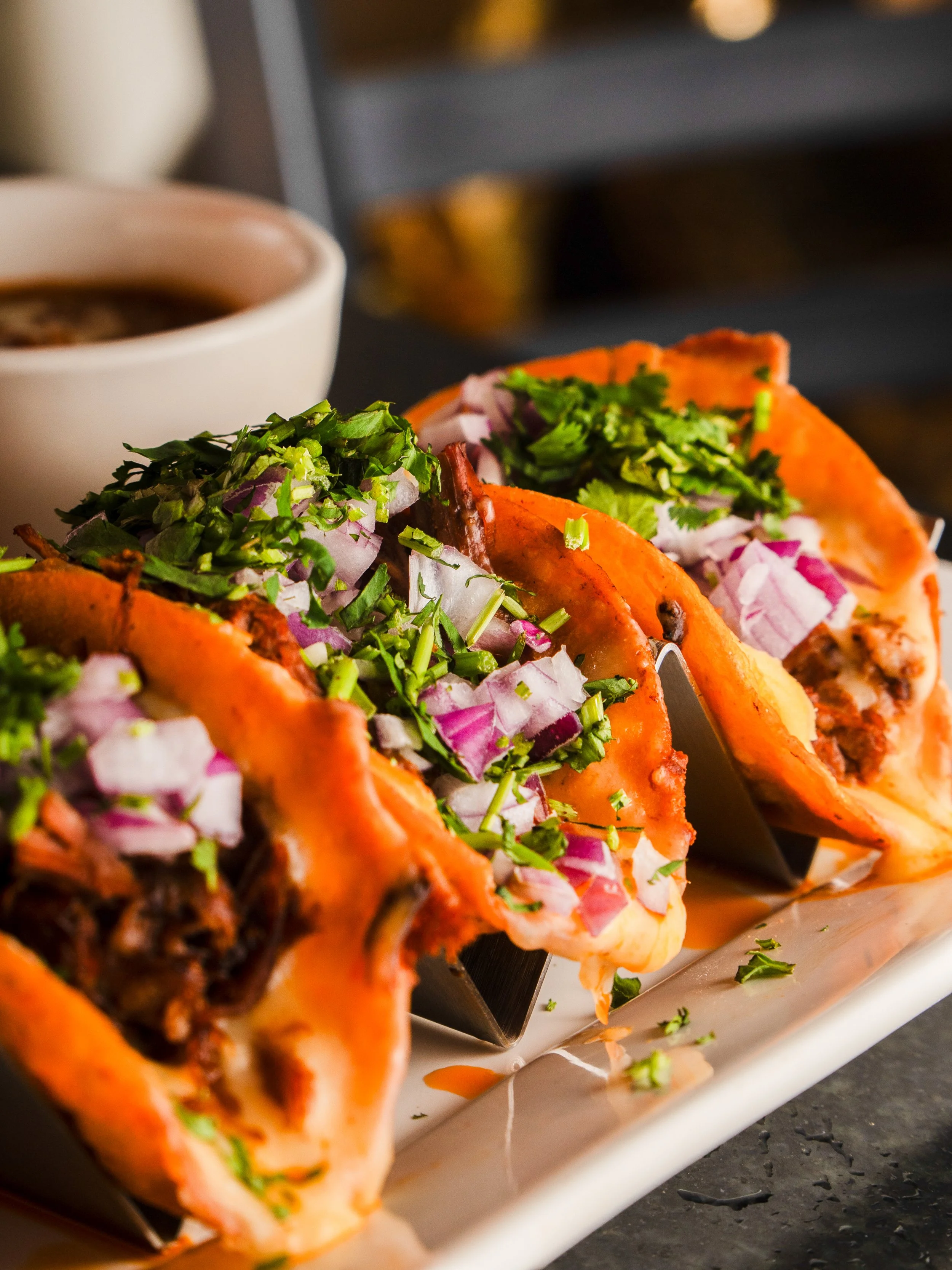 Close-up of three Birria tacos filled with meat, topped with chopped onions and cilantro, served on a white plate.