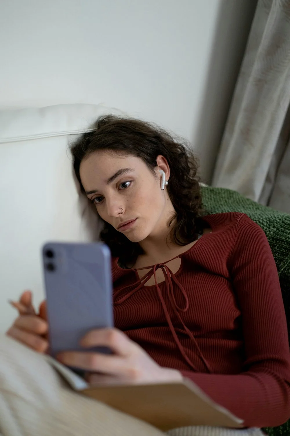Photograph of a young White woman looking at her phone, wearing wireless headphones, with a distant or sad look on her face. She has curly brown hair and pale skin.