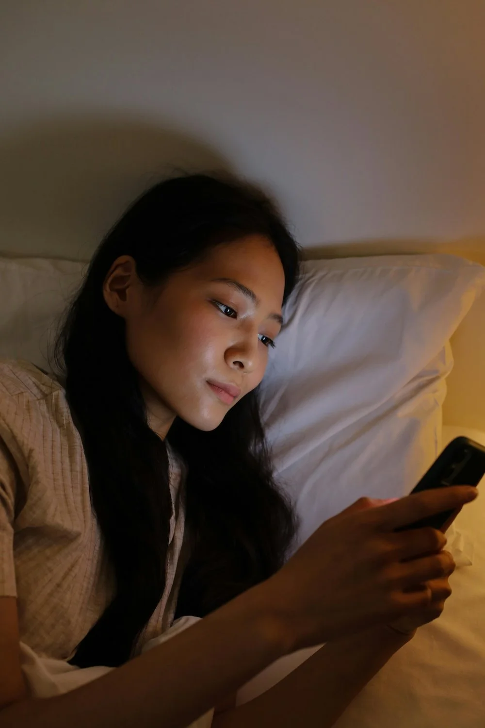 Photograph of a young East Asian woman looking at her phone in bed. She has long dark hair and light brown skin and looks serious.