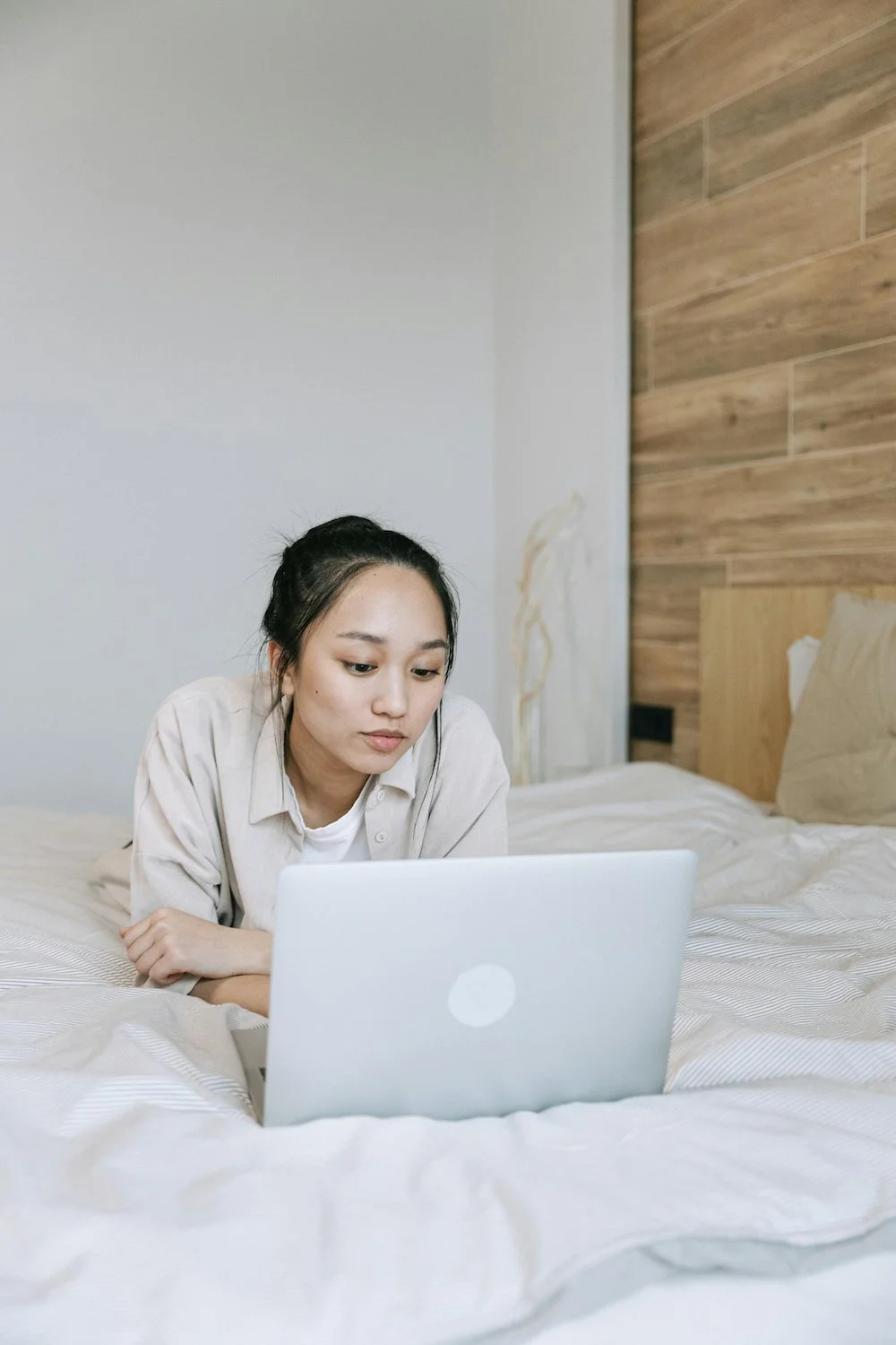 Photograph of a young East Asian woman with pale skin and dark hair looking at her laptop in bed.