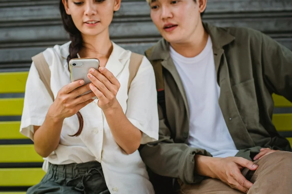 Photograph of a young East Asian couple looking at the woman's phone. They sit at a bus stop, both wearing backpacks, and have light skin and brown hair.