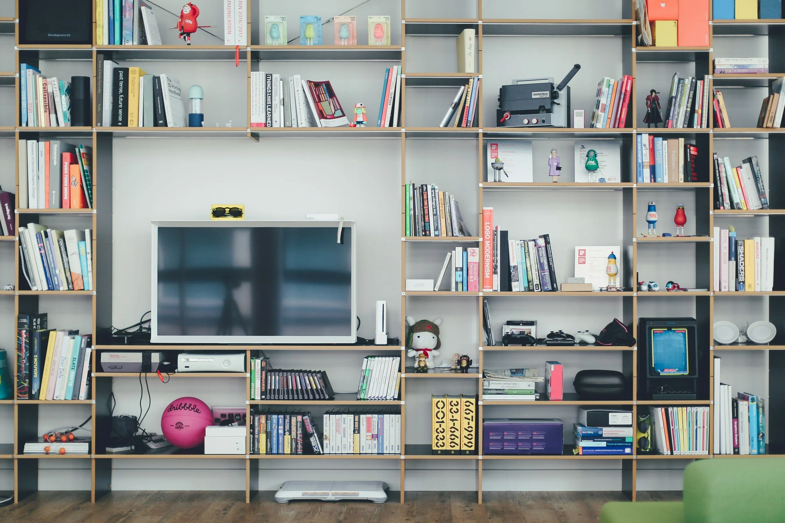 A large wooden bookshelf filled with books, toys, and decorative items. A flat-screen TV is mounted on the wall of the bookshelf, with a gaming console and other electronics below it. The bookcase has multiple compartments with various items including books, figurines, a pink exercise ball, and a vintage radio. The floor is wooden, and a green chair is partially visible in the bottom right corner.