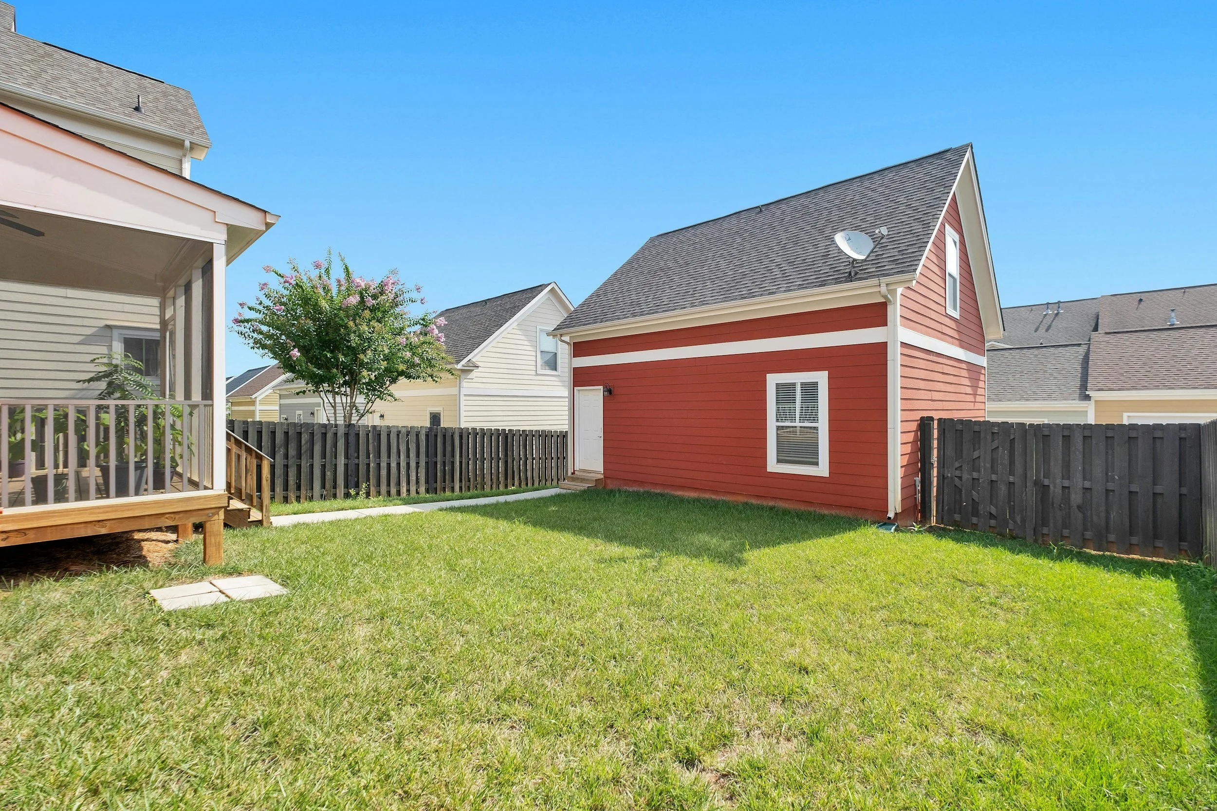 Backyard with a grassy lawn, a red shed with white trim, a black wooden fence, a porch with railing on the left, and neighboring houses with gray roofs and white walls under a clear blue sky.