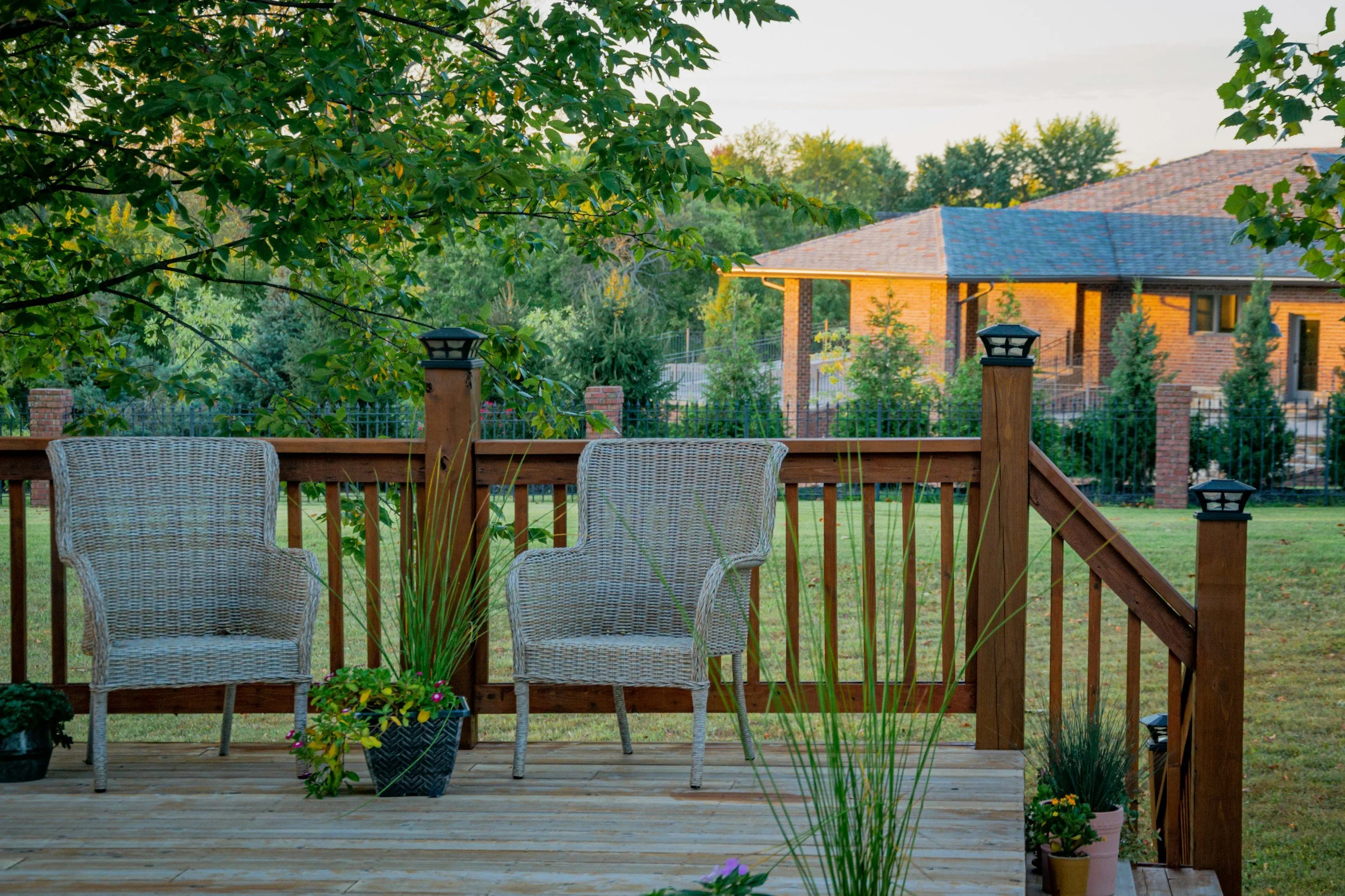 A wooden deck with two wicker chairs, potted plants, and a railing, overlooking a backyard with trees and a brick house in the background during daytime.