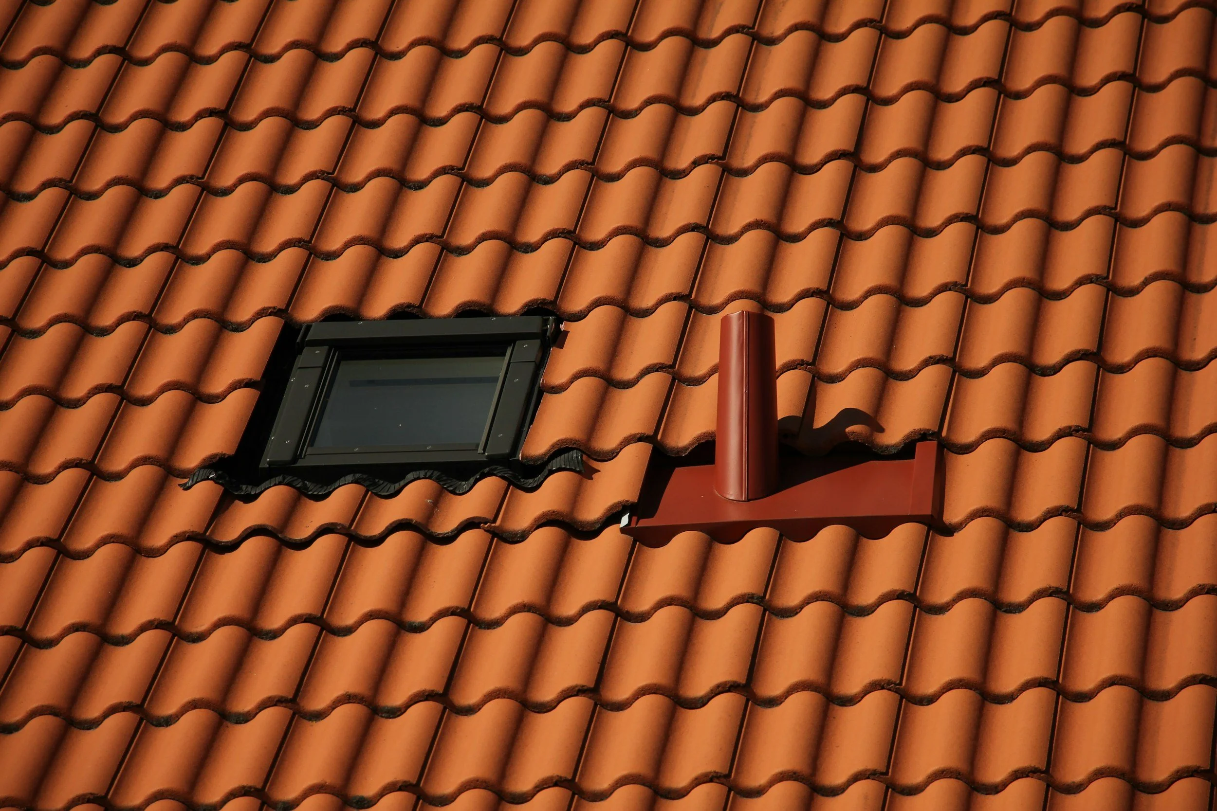 Close-up photo of a terracotta tiled roof with a small window and a red chimney vent.