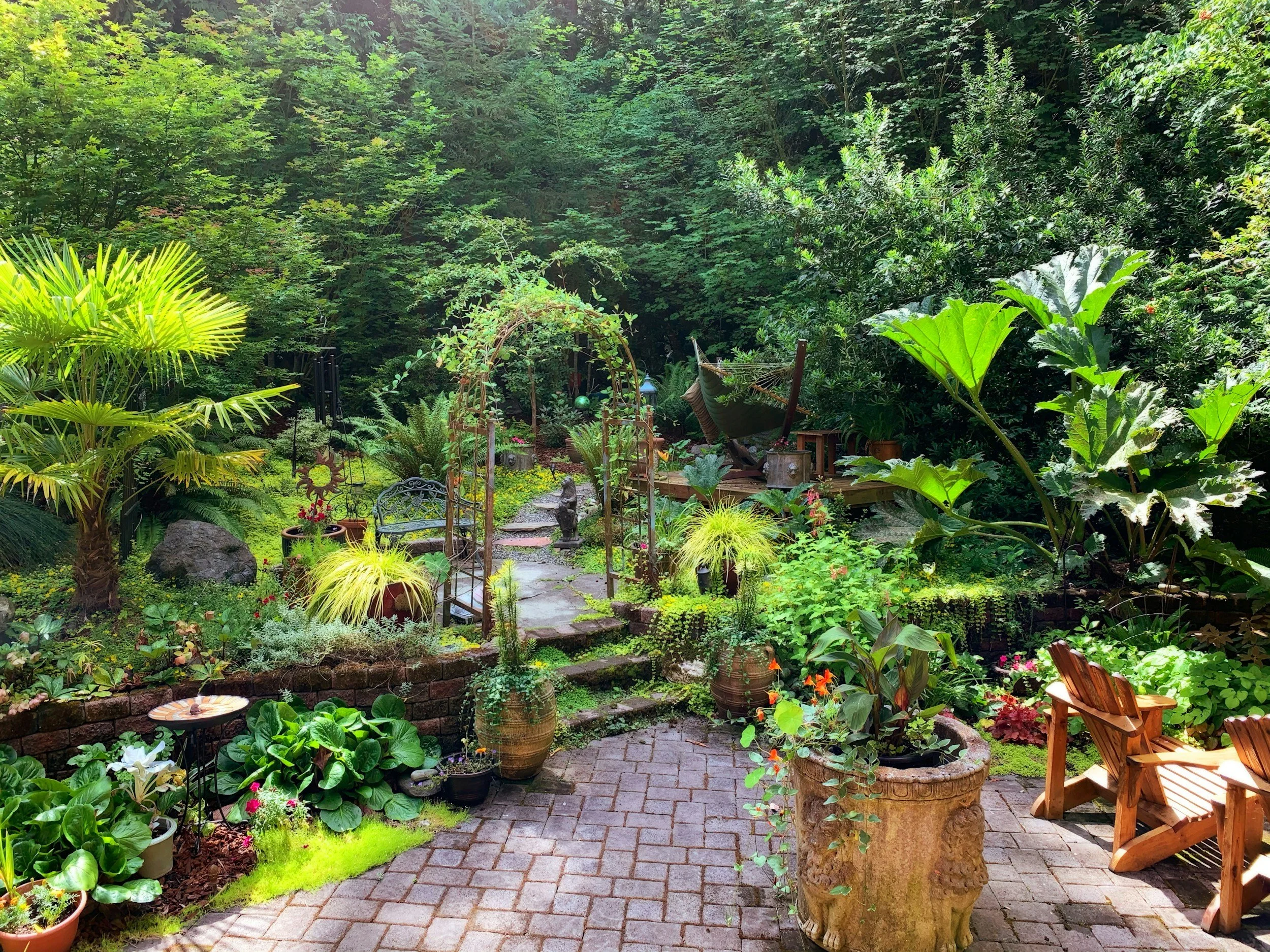 A lush garden with paved patio area featuring potted plants, garden furniture, and a brick border. A pathway leads into the dense, green forested background with various plants, trees, and decorative elements.