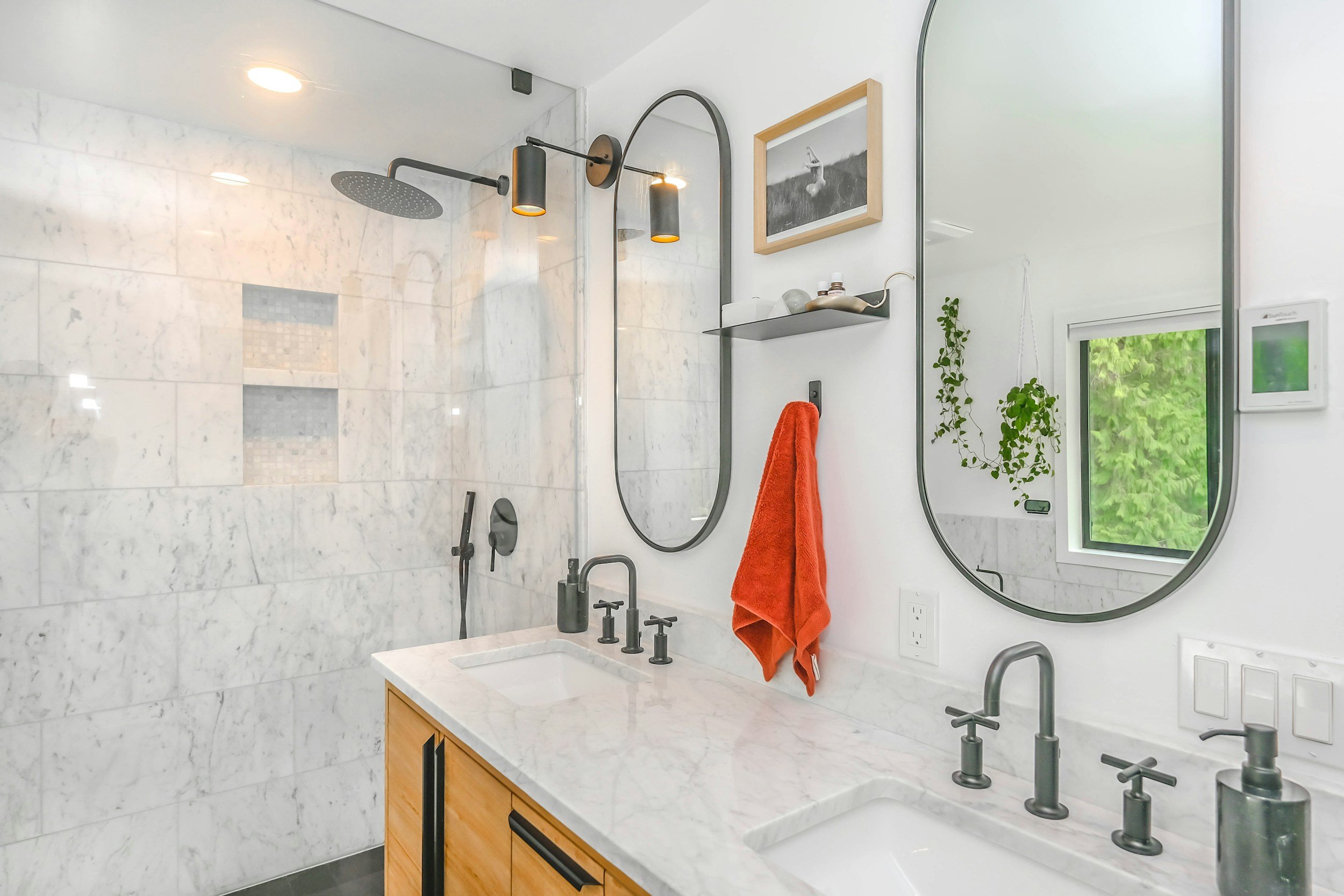 Modern bathroom with white marble walls and countertops, black fixtures, an orange towel, and round mirrors. There is a window with greenery outside and small plants hanging inside.