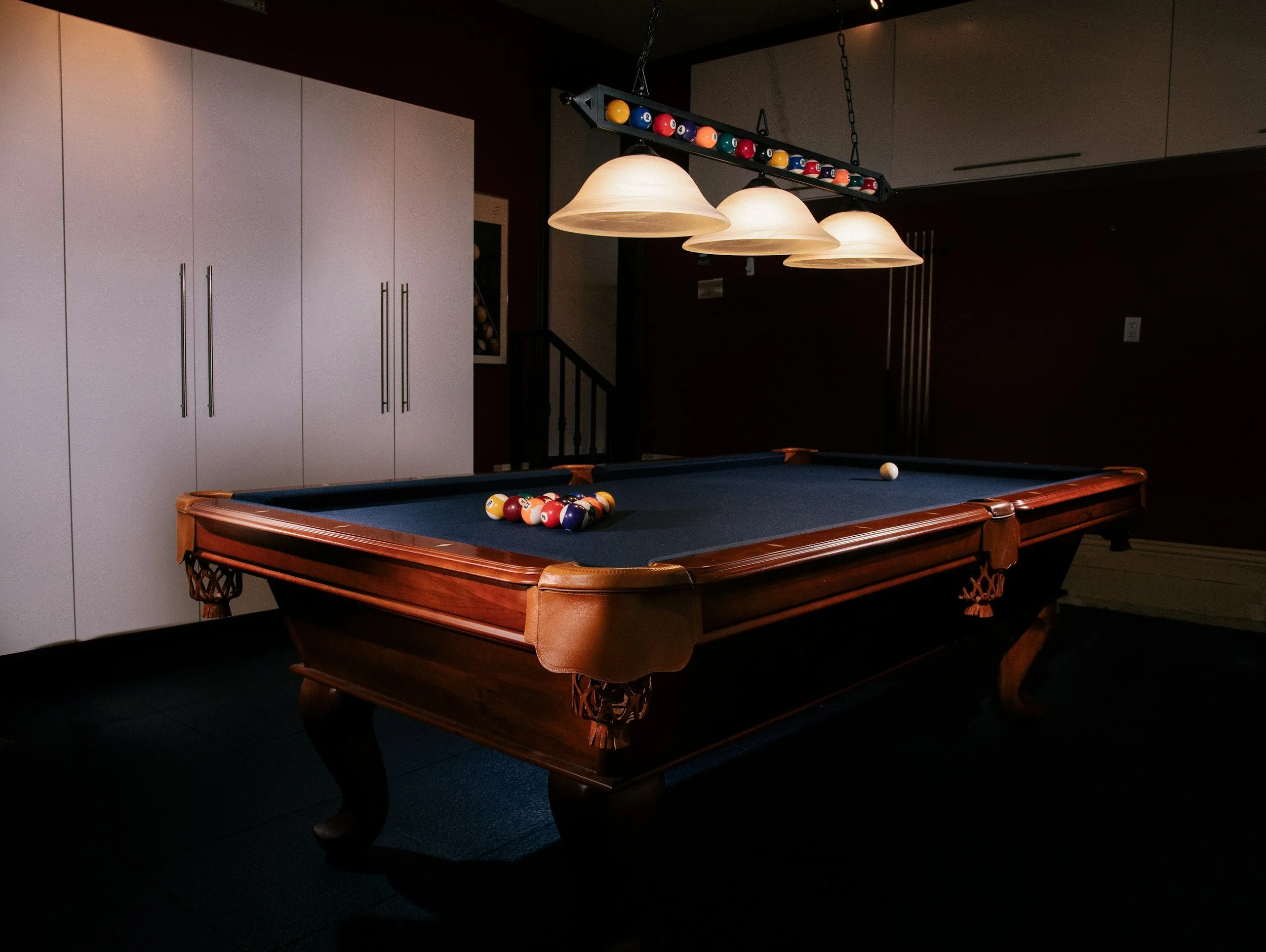 A pool table with a blue felt surface and a set of billiard balls racked at one end, in a dimly lit room with white cabinets and pendant lights hanging above.